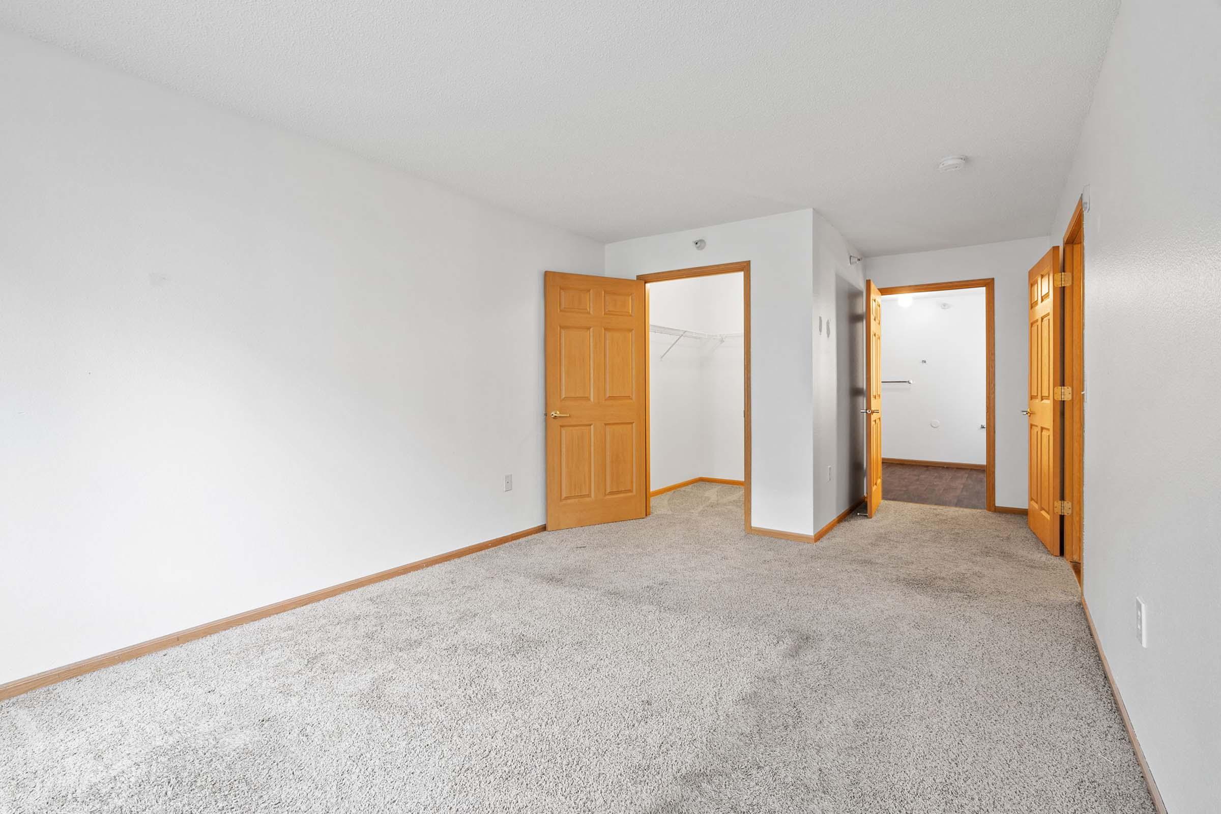 A spacious, empty room featuring beige carpet and white walls. Two wooden doors are visible, leading to a closet and another room. Natural light brightens the space, creating a clean and minimalistic atmosphere. No furniture or decor is present, emphasizing the room's potential for customization.