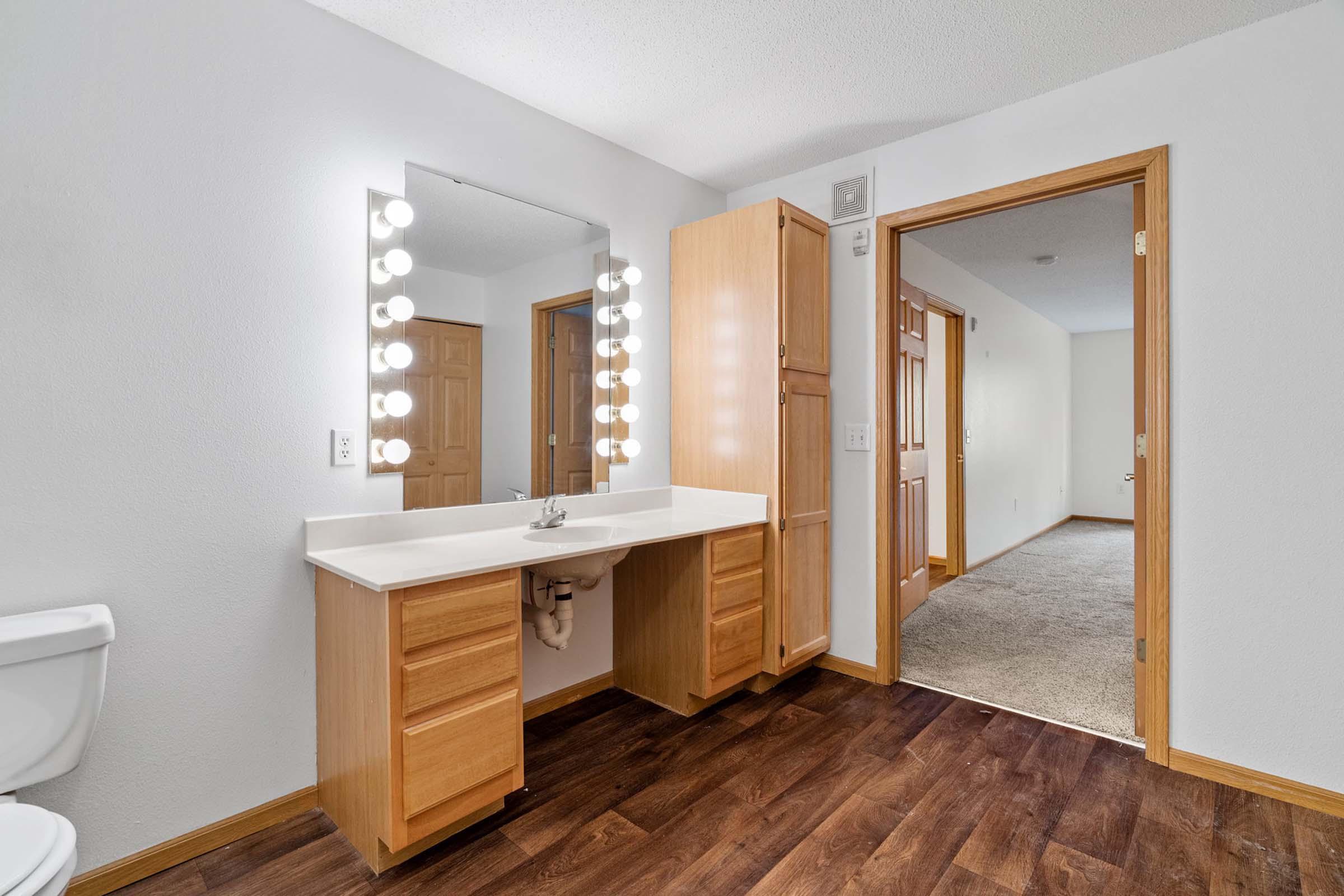 A well-lit bathroom featuring a modern vanity with a large mirror and bulbs, wooden cabinetry, a toilet, and a doorway leading to a carpeted area. The walls are painted light, and the flooring is dark wood, creating a warm, inviting atmosphere.