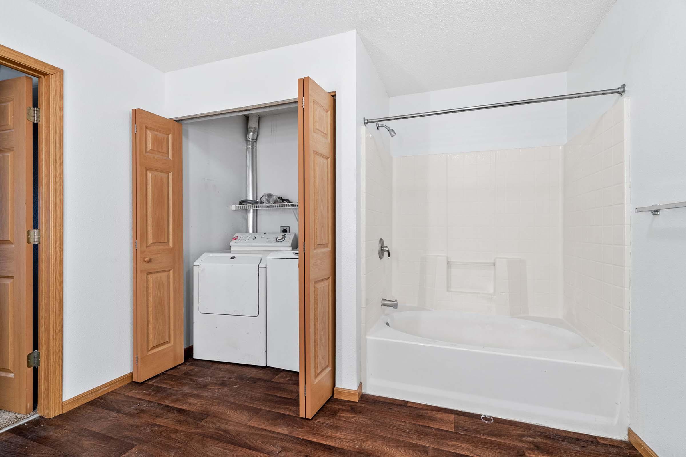 A small laundry area with a washer and dryer inside a closet, beside a bathroom featuring a bathtub and shower. The walls are painted white, and there are wooden doors and a dark wood floor.