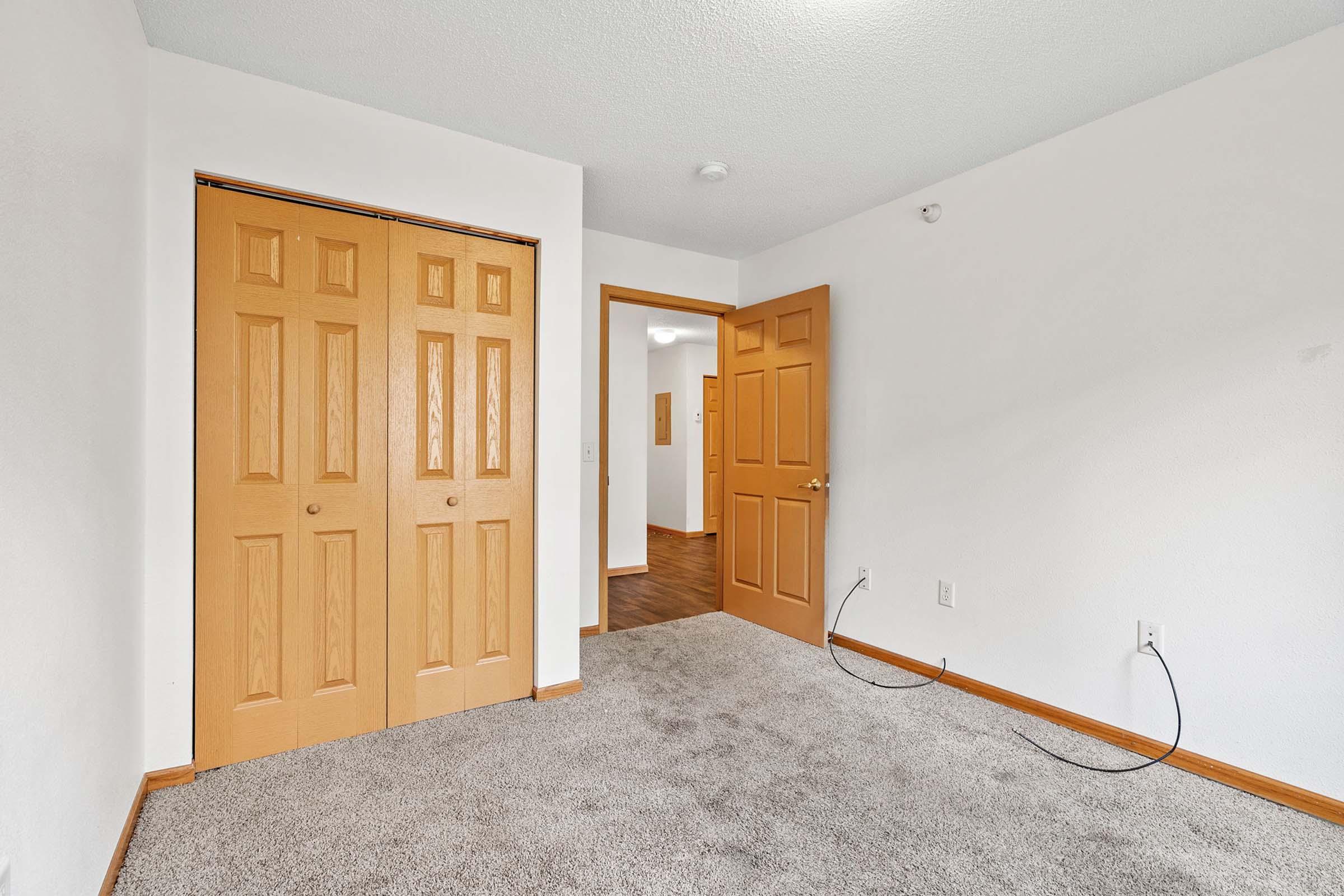 Empty room with light gray carpet, featuring a neutral-colored wall. There are two wooden double doors on the left, a doorway leading to another room on the right, and visible electrical wires on the floor. The overall space appears bright and unadorned, suggesting it is ready for furnishings.