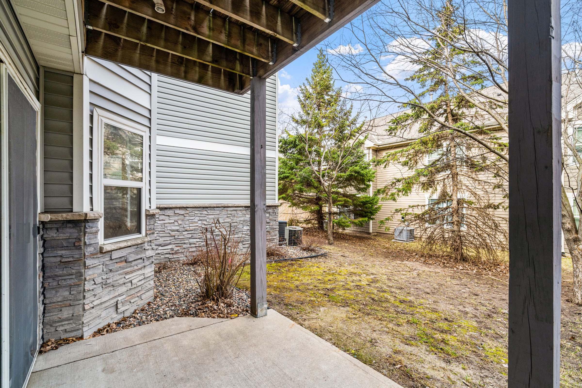 View from a covered porch showcasing a small outdoor area. The scene includes a mix of trees and shrubs, with a stone and siding wall structure nearby. Air conditioning units are visible in the background, and the ground is mostly bare with patches of grass and some decorative gravel.
