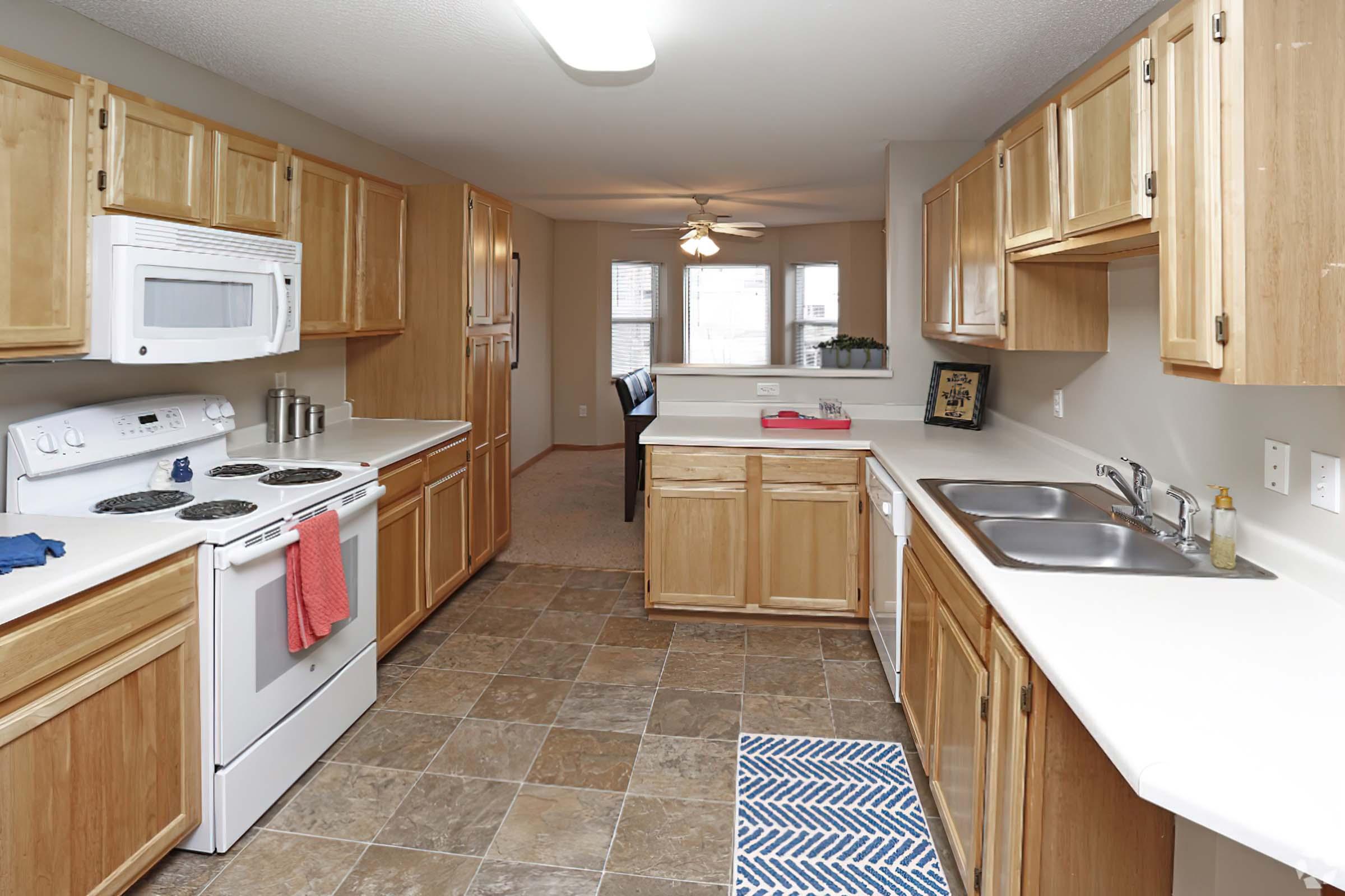 A modern kitchen featuring wooden cabinets, a white stove, microwave, and double sink. The countertops are light-colored, and a blue and white striped rug is placed on the floor. A ceiling fan can be seen, and there is natural light coming through windows in the background, leading to a living area.