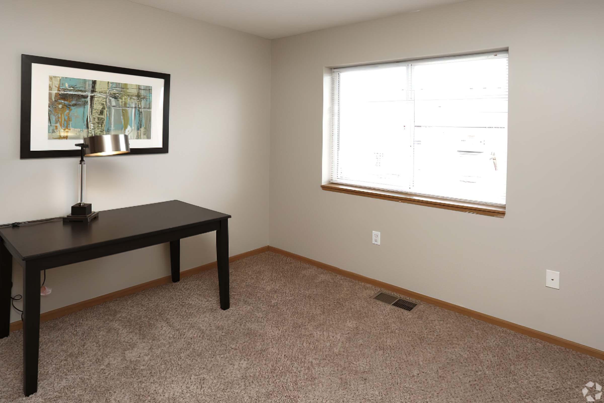 A minimalist room with light brown carpet, featuring a black desk with a lamp and a window with blinds. There is an abstract framed artwork on the wall and no furniture aside from the desk. The room is well-lit with natural light coming through the window.