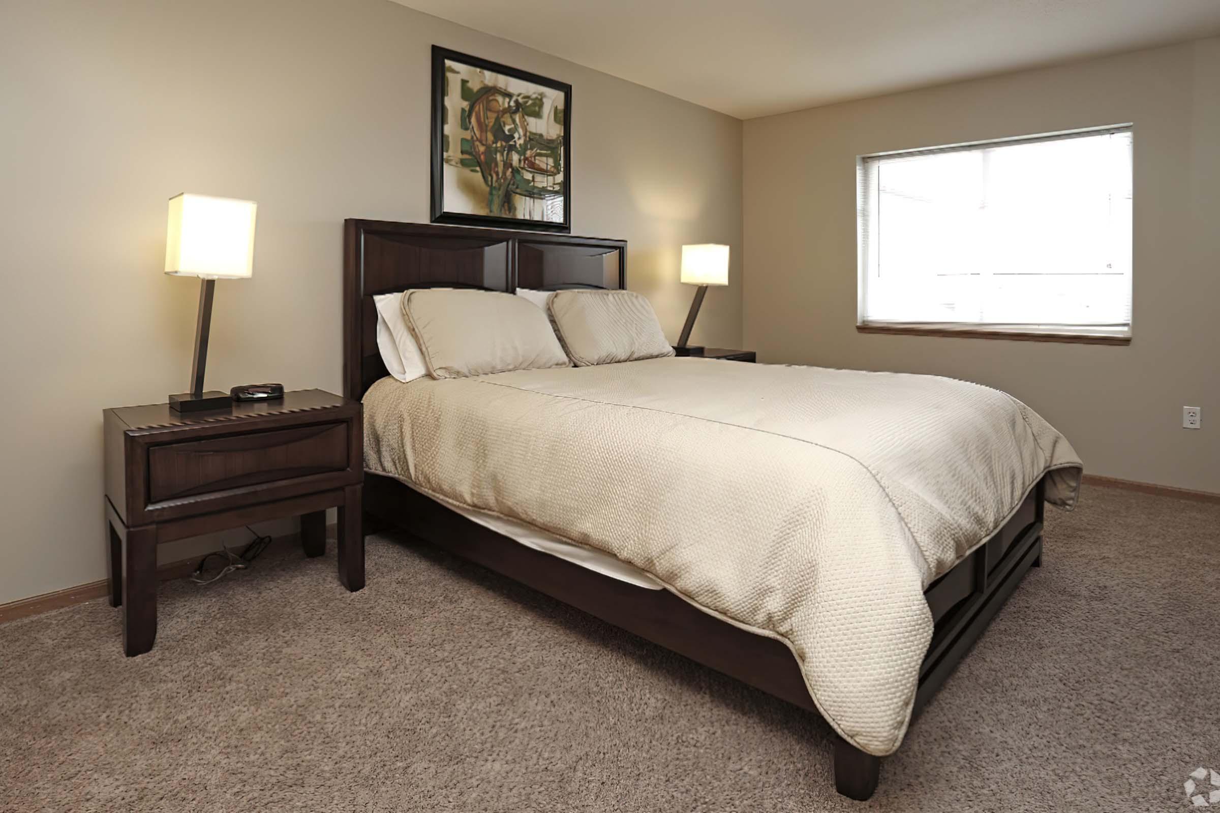 A well-lit bedroom featuring a queen-sized bed with a beige quilt, flanked by two bedside lamps on dark wood nightstands. A framed artwork hangs on the wall above the bed, and a window allows natural light to enter the space, which has neutral-colored walls and carpet.