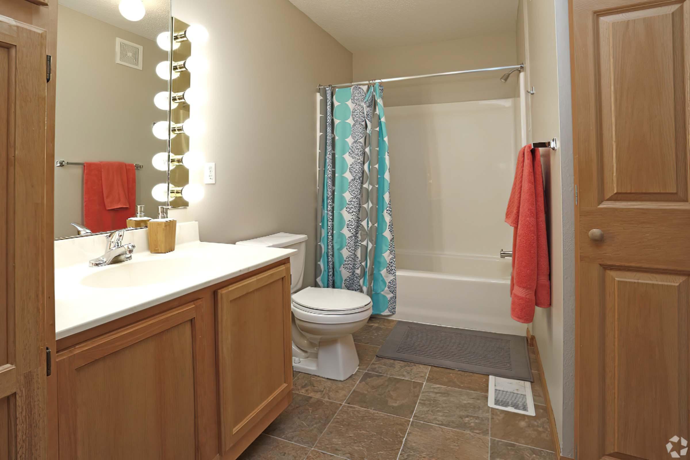 A clean and bright bathroom featuring a bathtub with a shower curtain, a vanity with a sink and wooden cabinets, and a toilet. The walls are light-colored, and there are colorful towels hanging. The floor is tiled, and there is a rug in front of the bathtub. Decorative lighting is above the mirror.