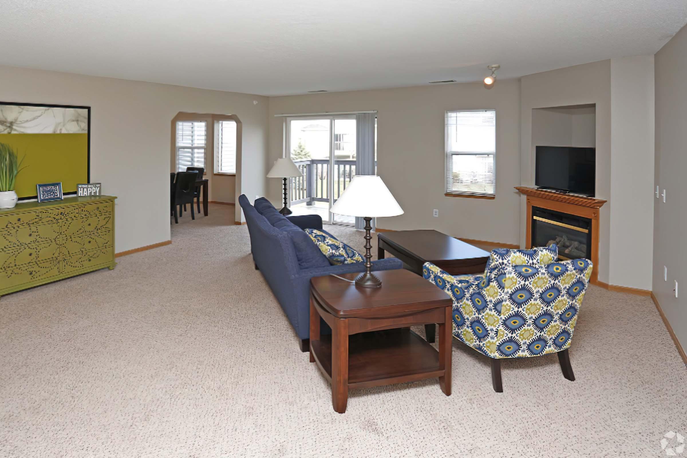 Spacious living room featuring a blue sofa and a patterned armchair, a wooden coffee table, and a lamp. A TV is placed on a stand near a cozy fireplace. Large windows provide natural light, and there is a view of a dining area in the background, with tasteful decor and neutral wall colors.