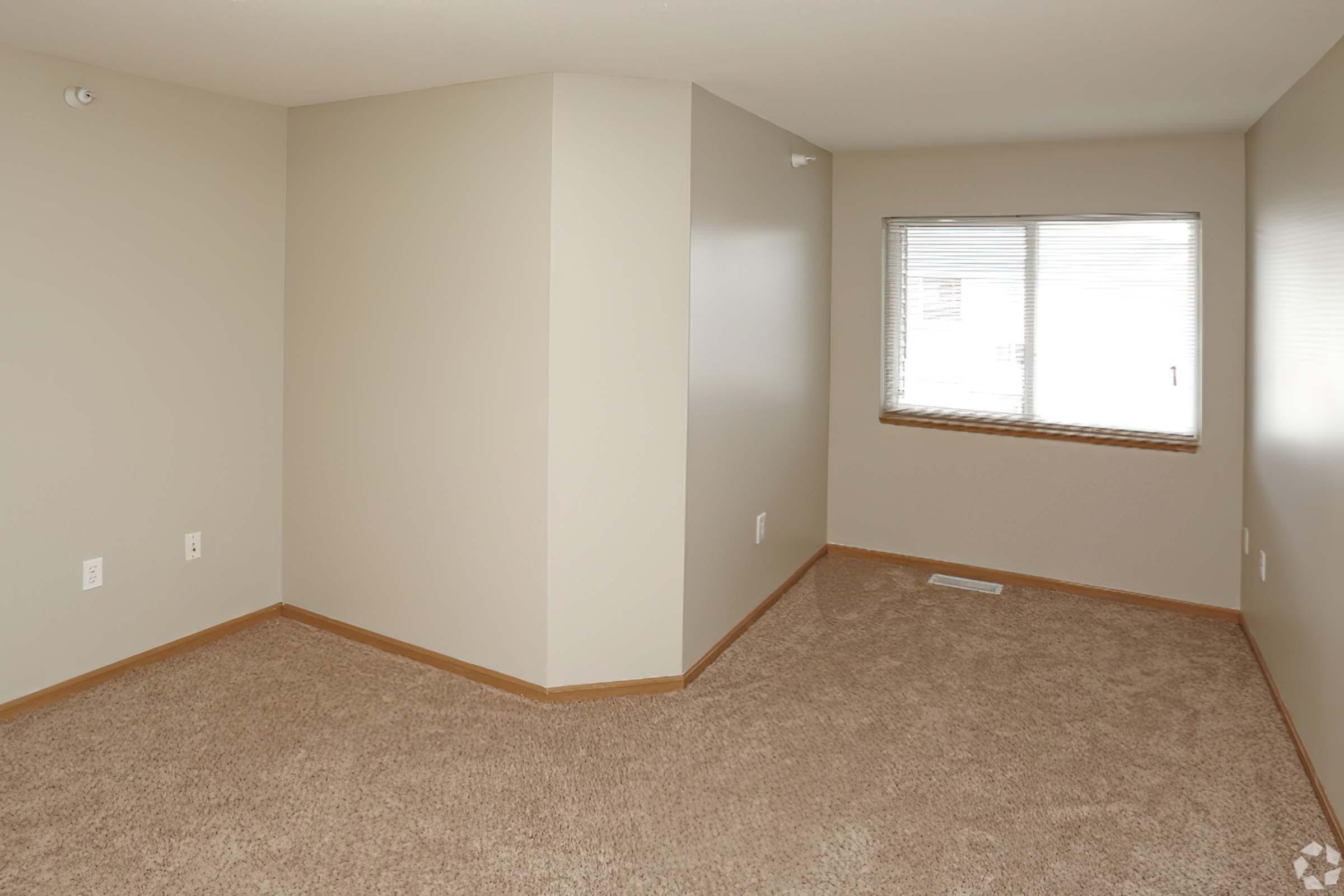 An empty, well-lit room featuring beige walls and carpeting. The space has a corner with a window that offers natural light, and the overall design is simple and modern, suitable for various uses.
