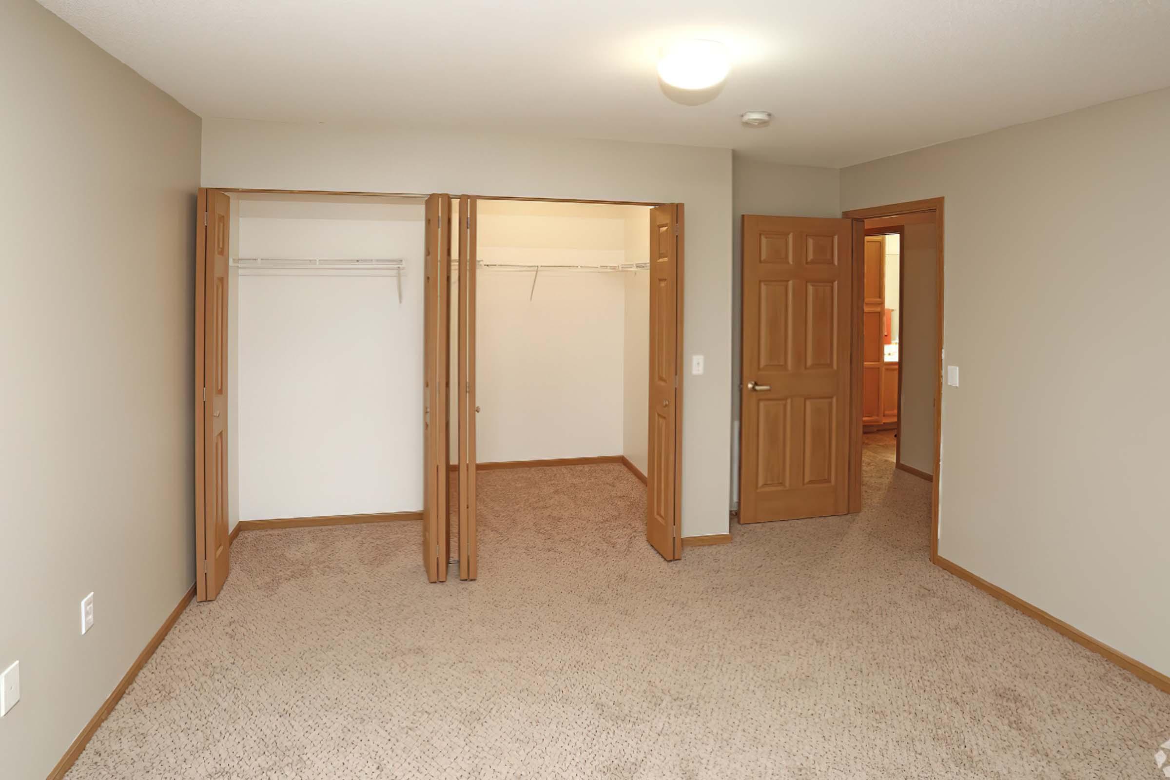 A spacious, empty bedroom with light beige walls and beige carpet. Two open closet doors on the left reveal empty storage space. A second door on the right leads to another room, and a ceiling light provides illumination. The overall atmosphere is bright and inviting.