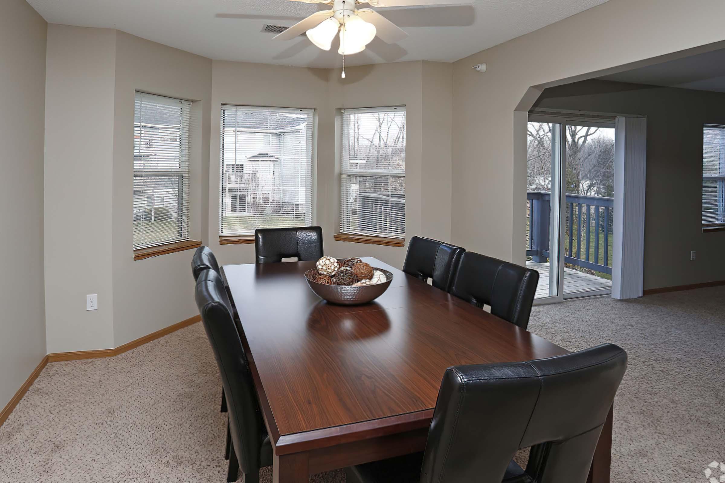 A spacious dining room featuring a large wooden table with a decorative bowl at the center, surrounded by black leather chairs. Large windows let in natural light, offering views of the outdoors, and the room has a cozy carpeted floor with neutral-colored walls and a ceiling fan.