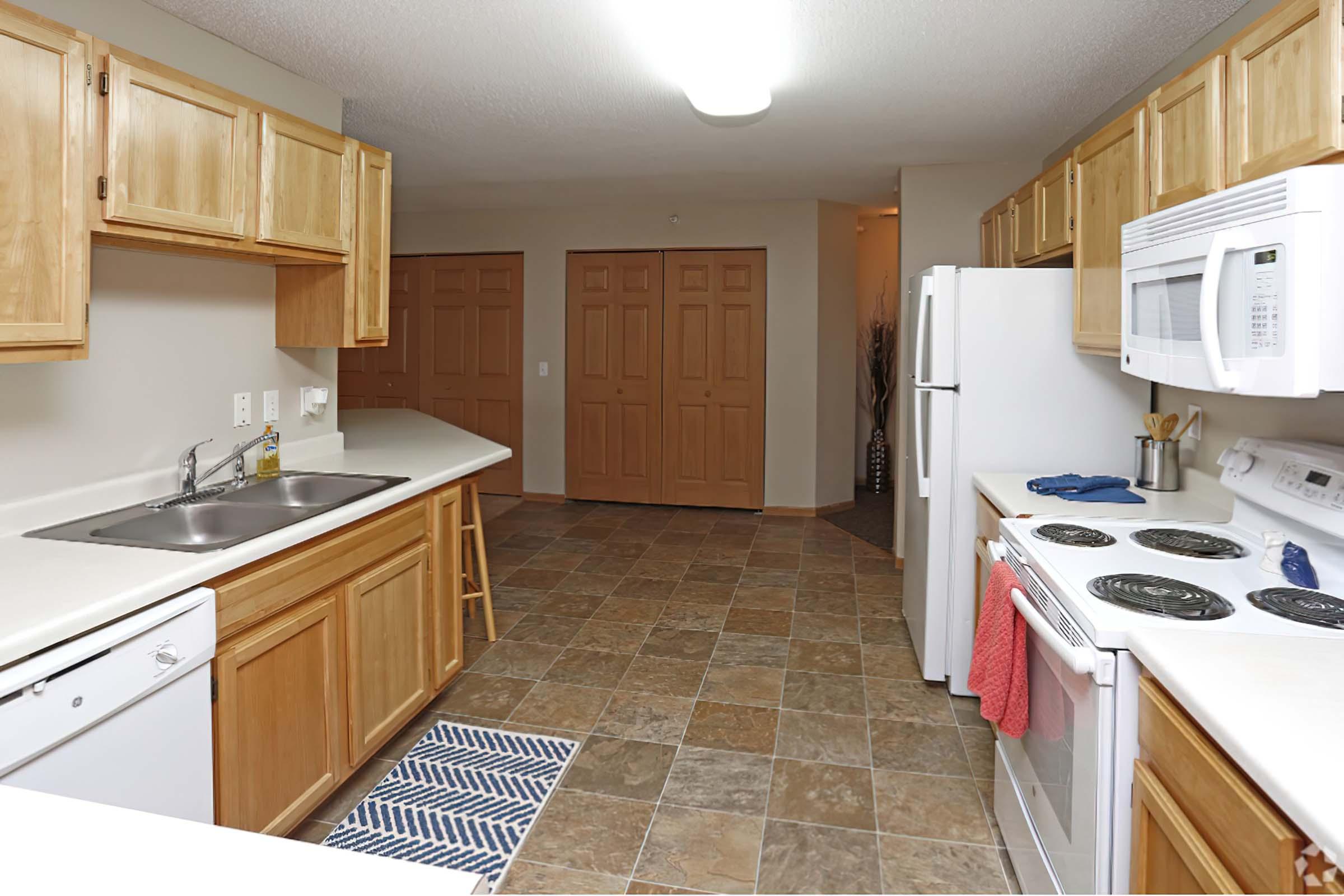 A modern kitchen featuring light wooden cabinets, a double basin sink, a dishwasher, and a stove with an oven. The countertops are white, and there’s a small bar stool by the sink. The floor has a tiled design, and a blue and white striped rug is placed in front of the sink. Doorways lead to another area in the background.