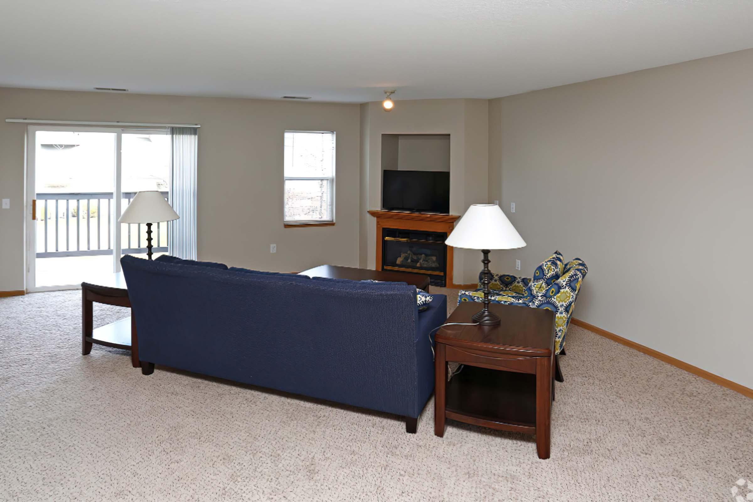 A spacious living room featuring a blue couch, a patterned armchair, a coffee table, and two lamps. Large windows allow natural light, with a sliding door leading to a patio. A TV is mounted on the wall above a wooden fireplace, and the room has neutral-colored walls and carpet.