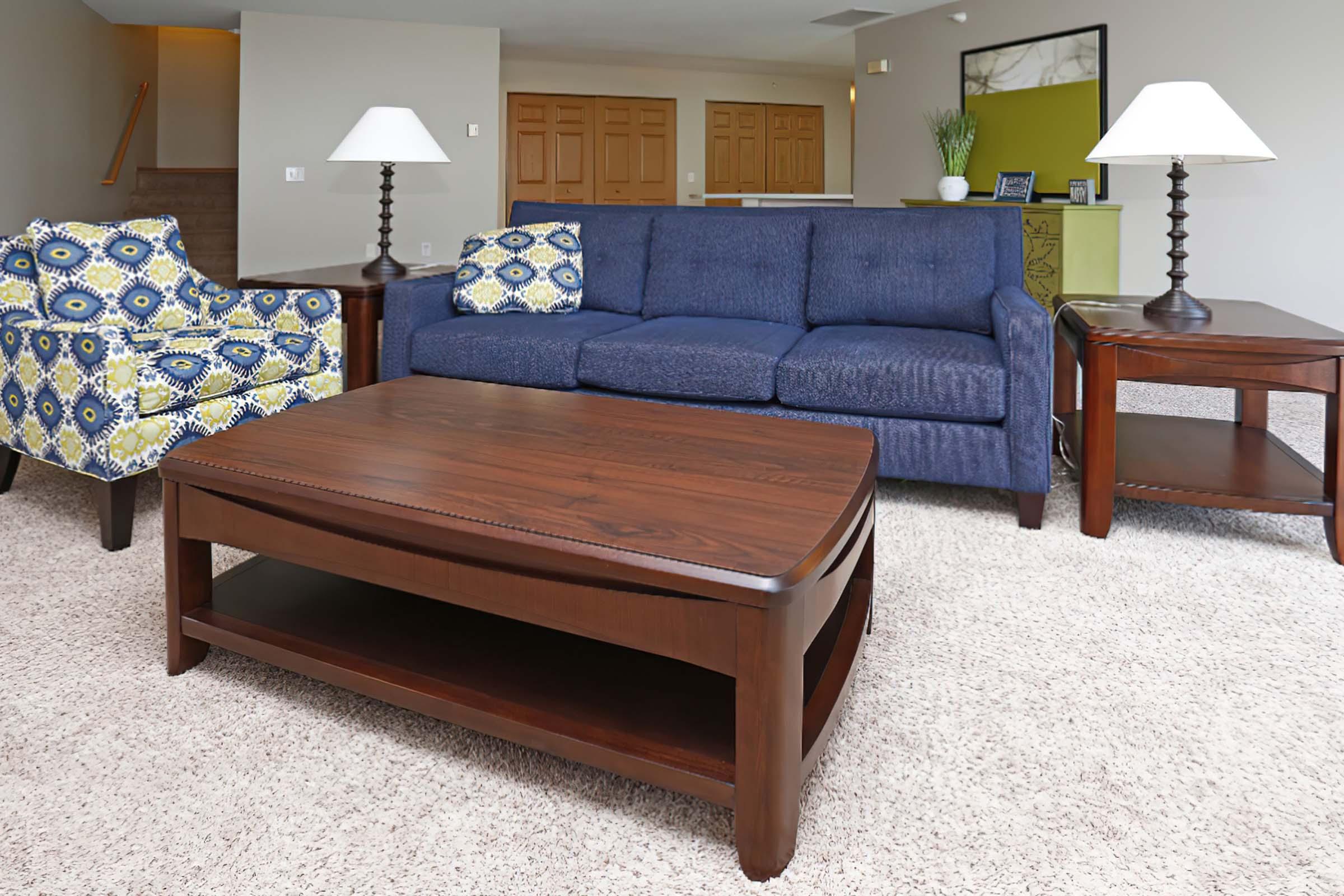 Living room setting featuring a blue sofa, patterned armchair with yellow and blue accents, and dark wood tables. The carpeted floor adds warmth, and lamps provide soft lighting. A staircase is visible in the background, along with a large window and a hint of a kitchen area.