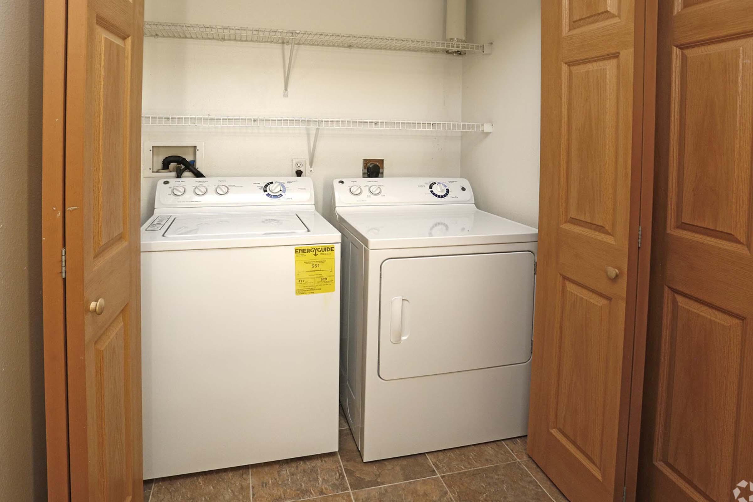 A laundry room featuring a white washing machine and a white dryer side by side. The appliances are set against a light-colored wall, and there are wooden closet doors partially closed. A shelf above the machines is empty, and the floor is tiled in a neutral color.