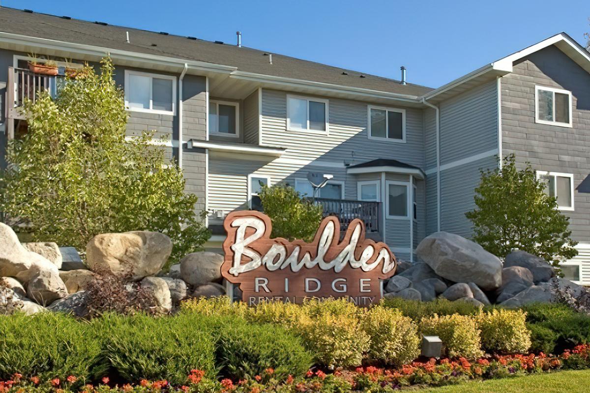 Sign for Boulder Ridge Rental Community, located in front of a multi-story residential building. The sign is surrounded by landscaped greenery, including shrubs and flowers, with large decorative rocks. The sky is clear and blue.