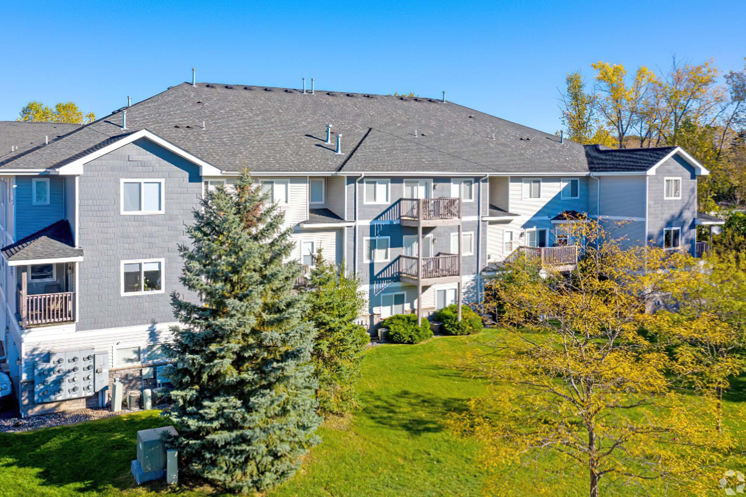 A multi-story residential building with a gray exterior and balconies, surrounded by lush green grass and a few trees. Clear blue sky overhead highlights the well-maintained landscaping and outdoor space.