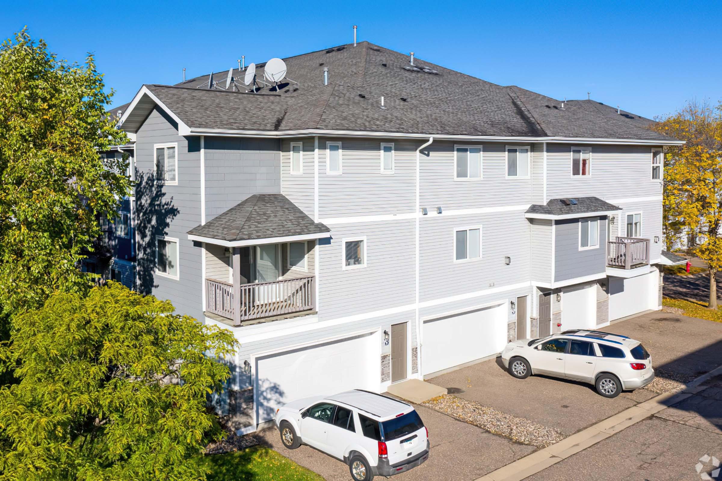 A row of modern townhouses with light gray siding and multiple balconies. The buildings feature white garage doors and outdoor parking spaces, surrounded by trees with green foliage. Clear blue sky overhead indicates a sunny day.