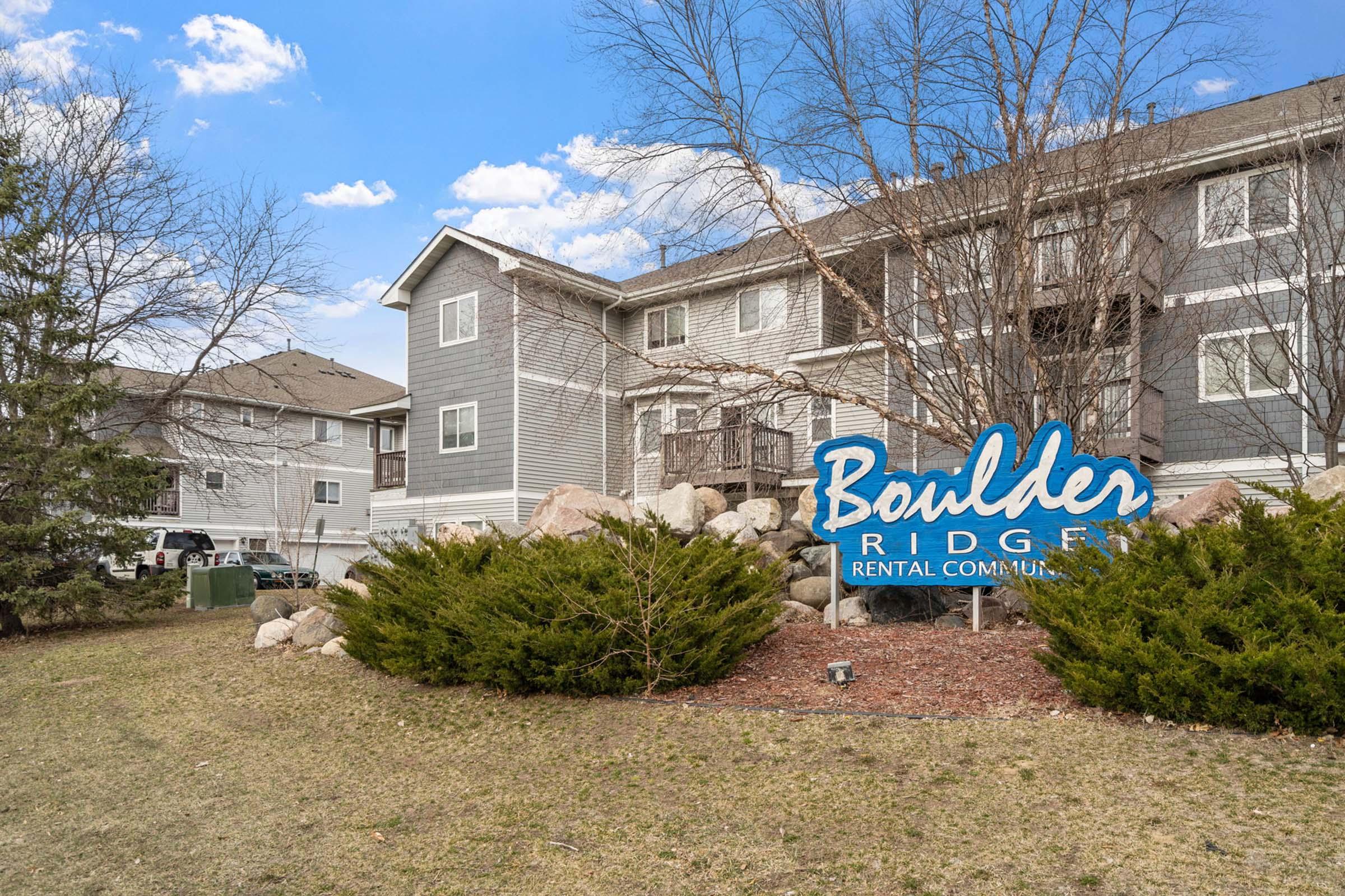 Sign for Boulder Ridge Rental Community, featuring blue lettering, surrounded by landscaped greenery and rocks. In the background, there are multiple residential buildings under a clear blue sky with scattered clouds.