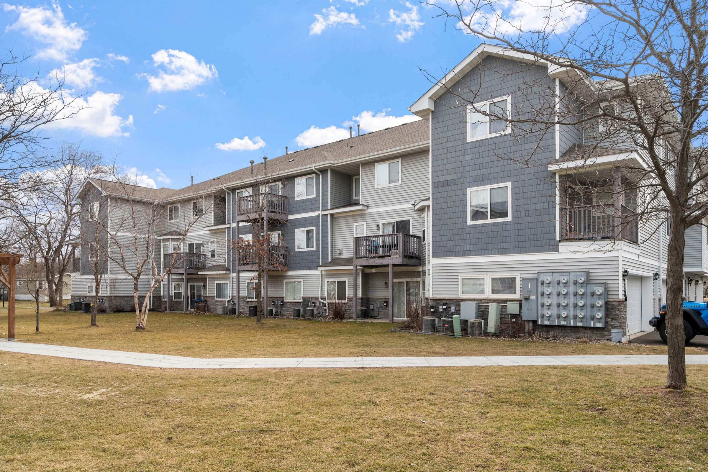 A multi-unit residential building featuring gray and beige siding, balconies, and multiple utility meters. The image shows a grassy area in front, with trees and a sidewalk, under a partly cloudy blue sky.