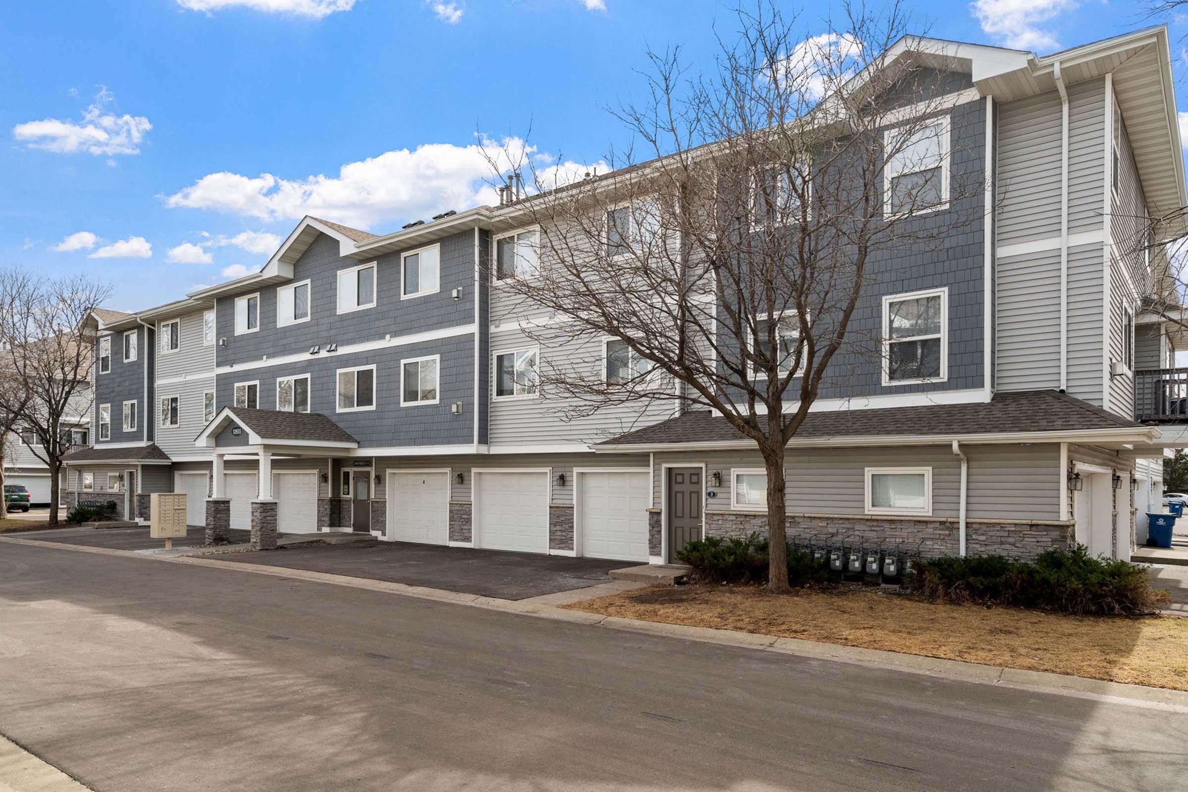 A multi-story residential building with a gray exterior, featuring a combination of siding and stone elements. The building has a row of garages and is flanked by empty parking spaces. Trees are visible in front, and the sky is clear with a few clouds.