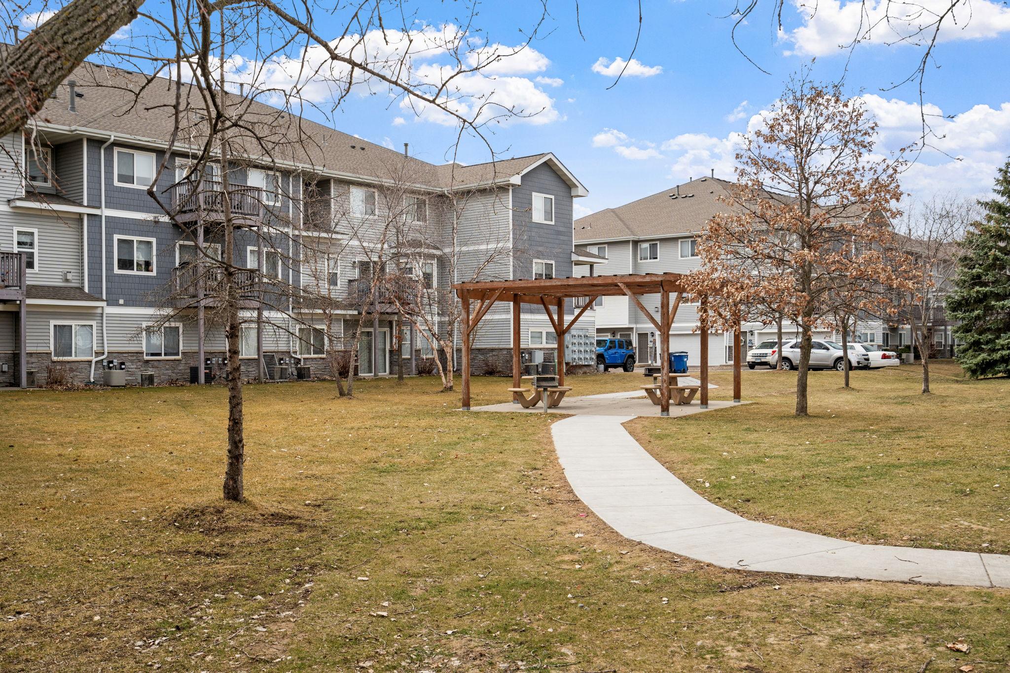 A grassy area featuring a paved walkway leading towards a wooden pergola with picnic tables underneath. Surrounding the area are two-story residential buildings, some trees, and parking spaces in the background, all under a partly cloudy sky.