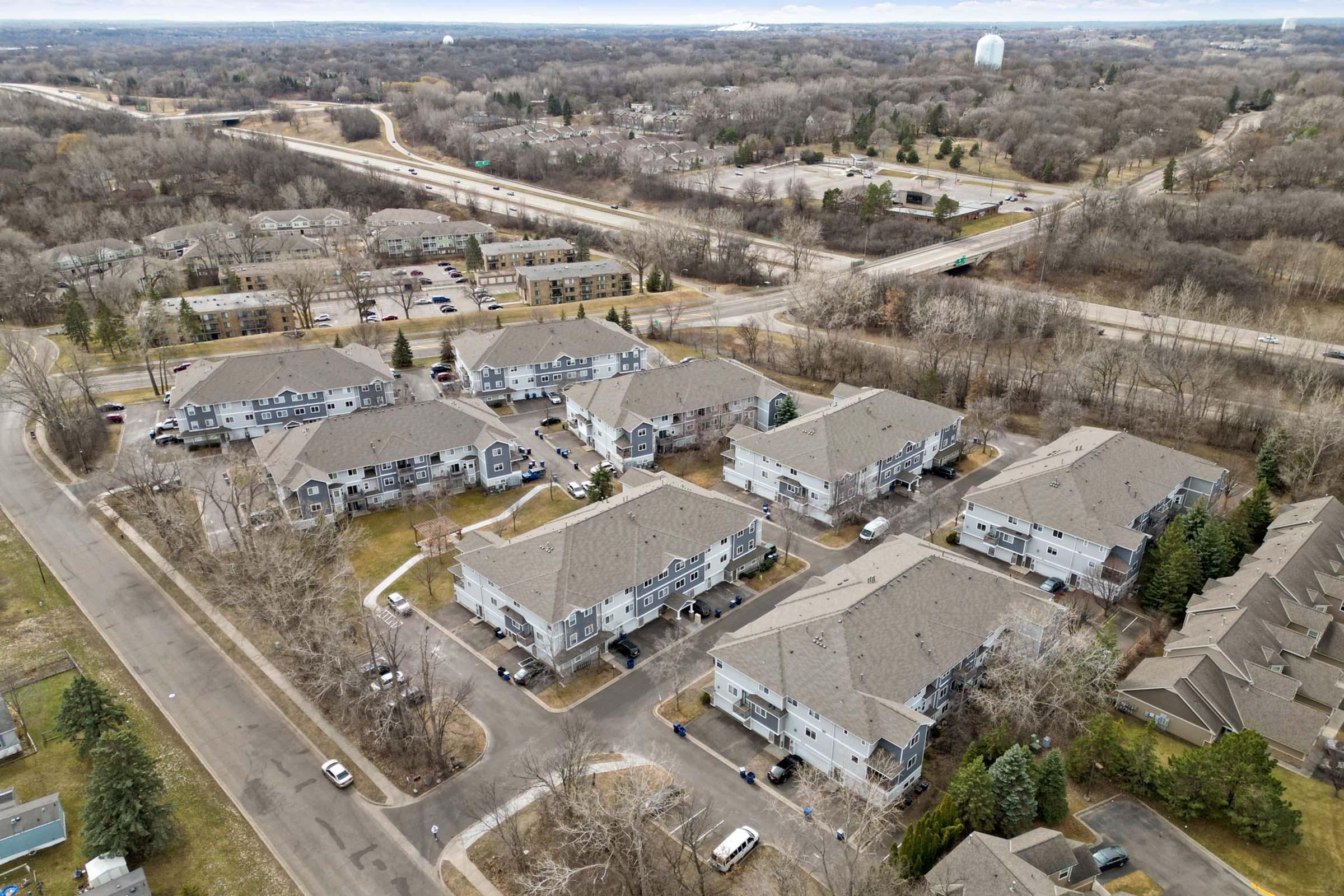 Aerial view of a residential area featuring multiple apartment buildings, surrounded by trees and open land. Nearby, a highway and other developments are visible in the distance, indicating a suburban setting. The scene captures a mix of green spaces and urban infrastructure.
