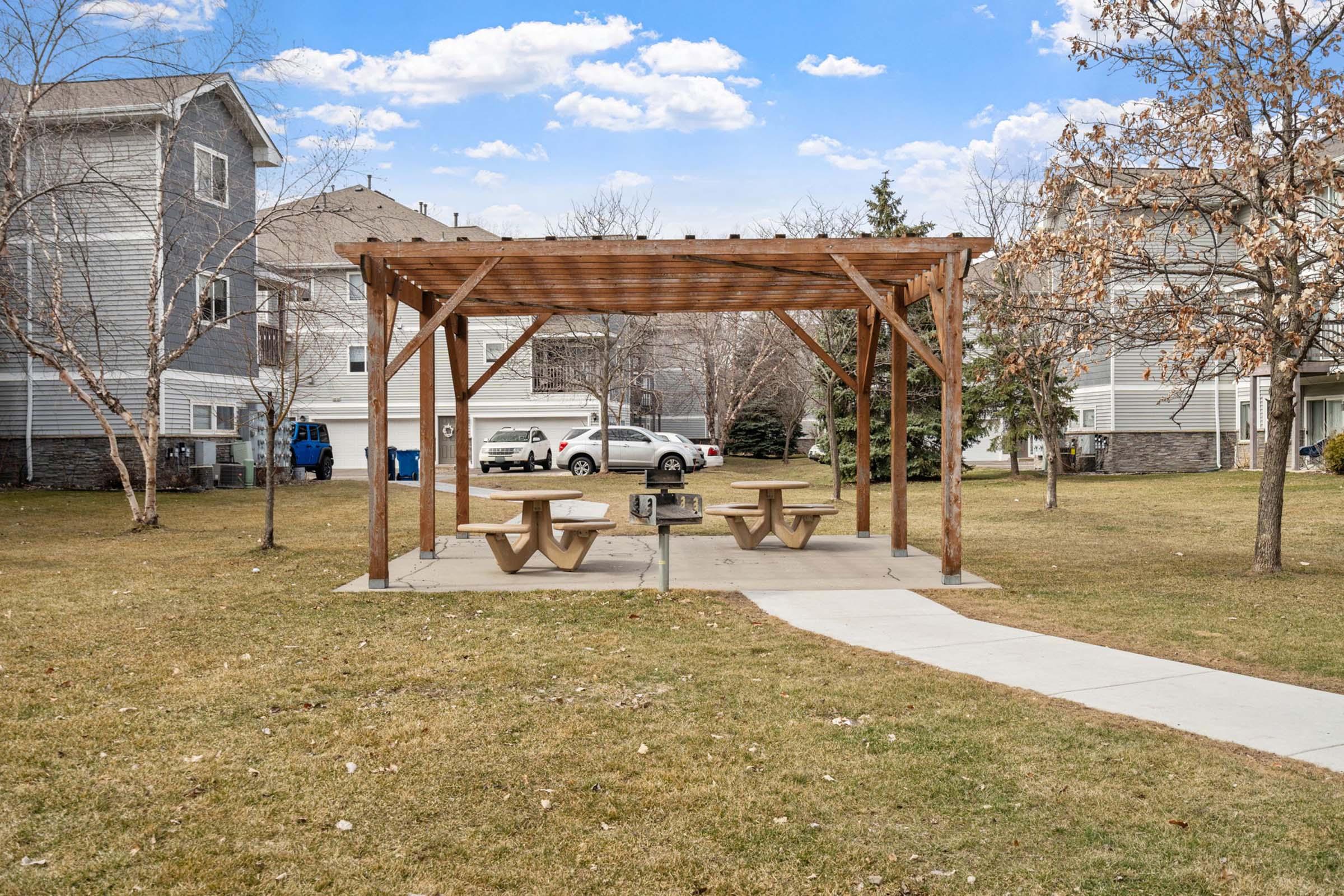 A wooden pergola over a picnic area with several tables on a grassy lawn, surrounded by trees and residential buildings in the background. The sky is partly cloudy, and there are parked cars near the homes. A pathway leads to the picnic area.