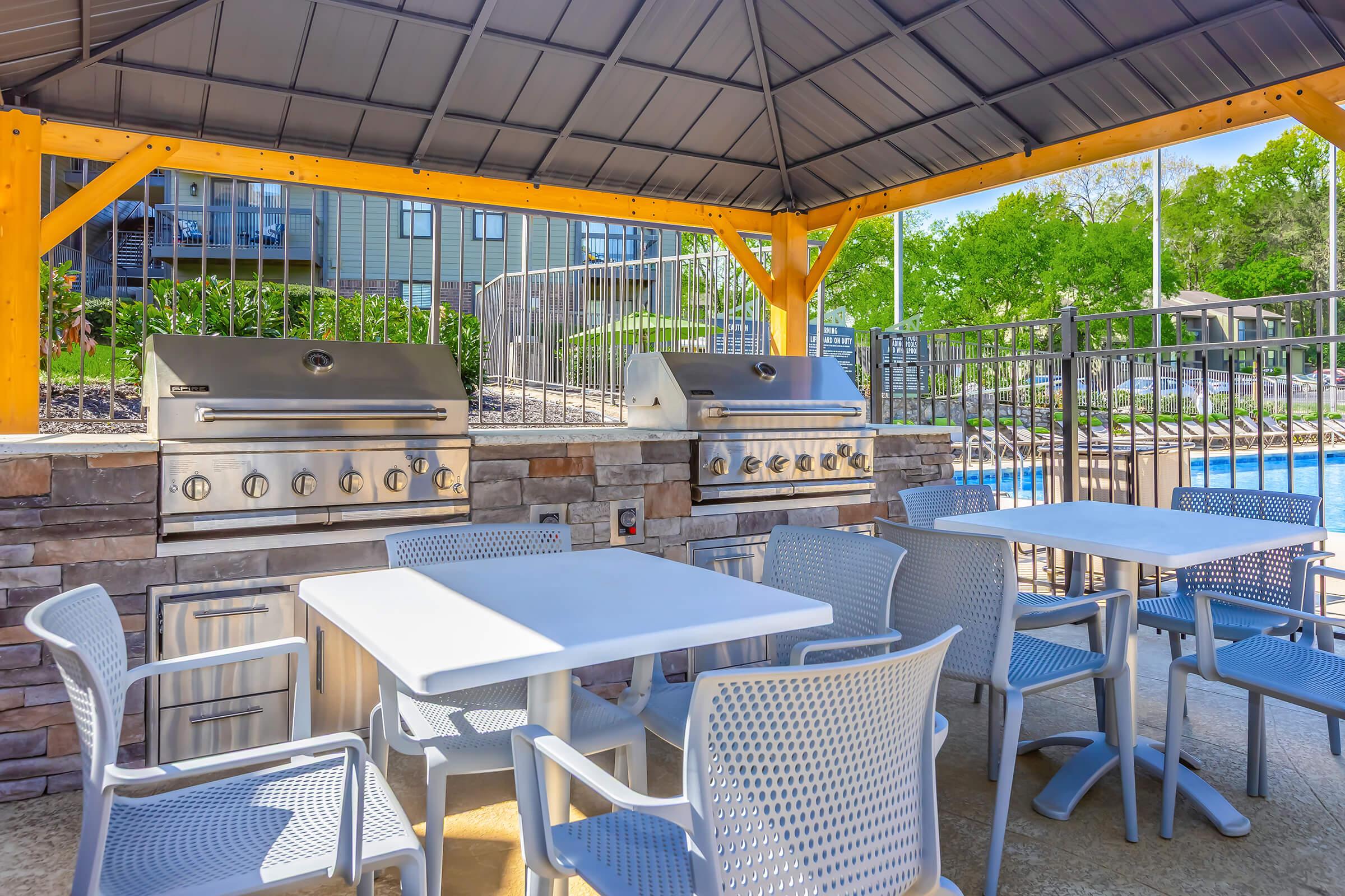 An outdoor grilling area featuring two stainless steel barbecue grills under a wooden gazebo. There are multiple white outdoor tables and chairs nearby, and the setting includes a well-manicured lawn and a swimming pool in the background. Bright greenery surrounds the area, creating a welcoming atmosphere.