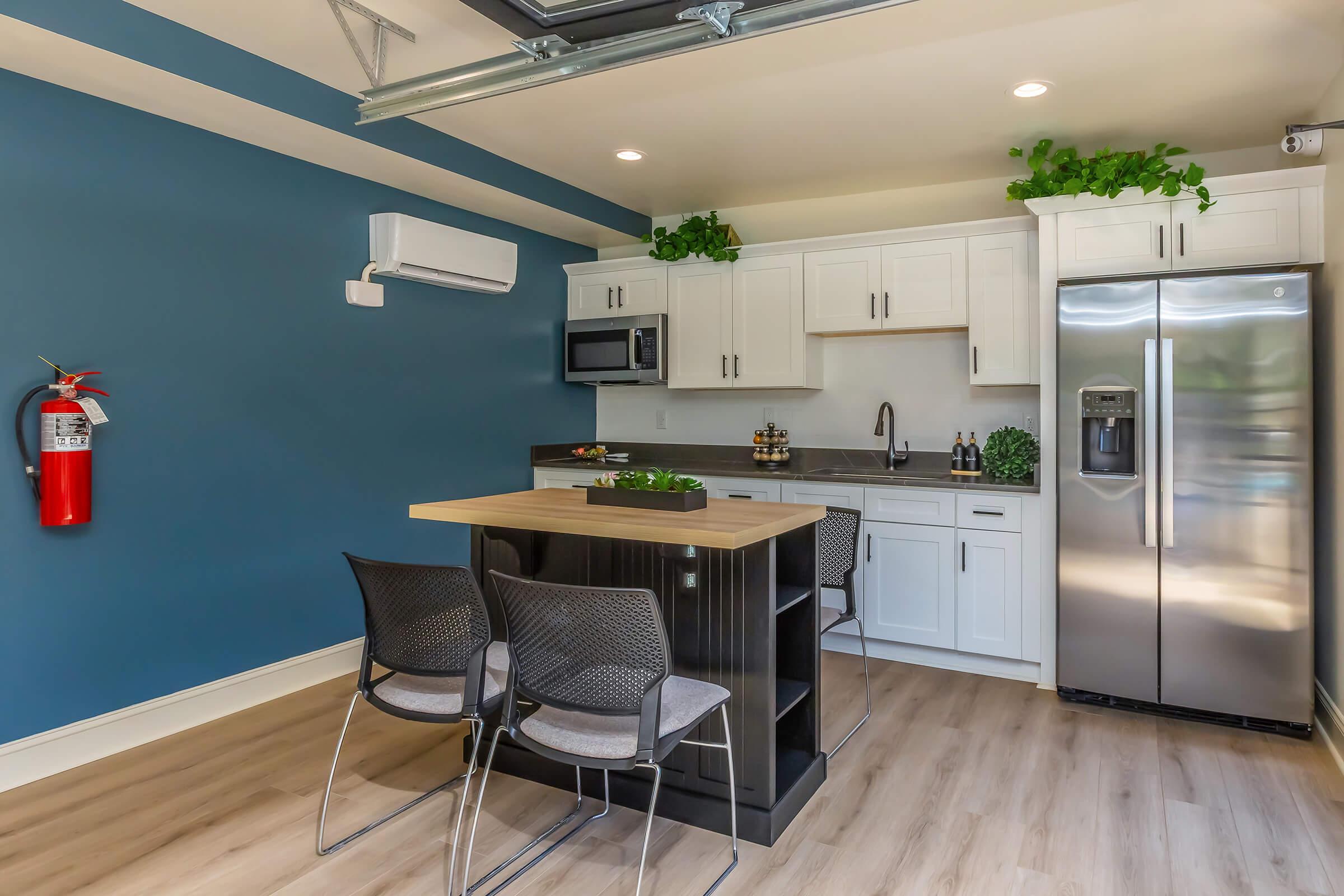Modern kitchen featuring white cabinets, stainless steel refrigerator, and microwave. A central island with a wooden top is surrounded by three black chairs. The walls are painted blue, and there are plants on the counter and shelves. A fire extinguisher is mounted on the wall. Bright, inviting atmosphere with wooden flooring.