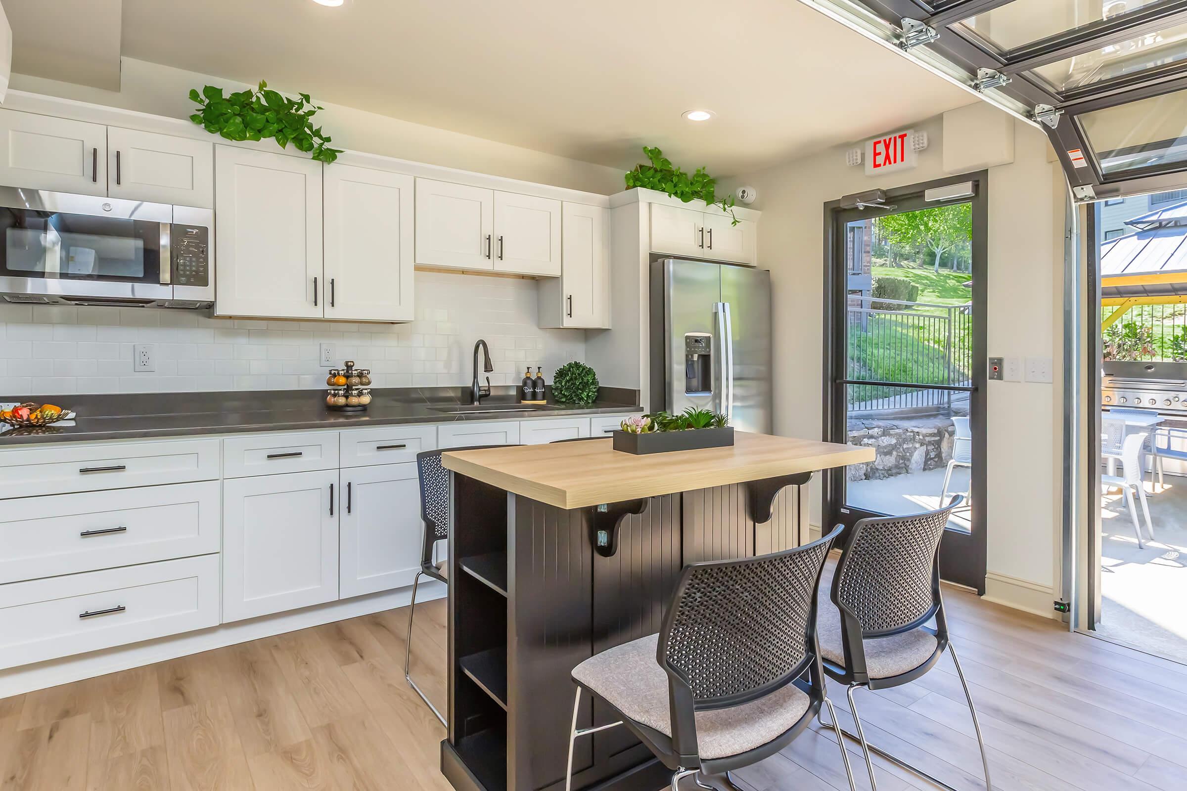 A modern kitchen featuring white cabinetry, a gray countertop, and stainless steel appliances. There's a central island with a wooden top and storage, along with four gray chairs. The space is well-lit with natural light coming through a large glass door that leads to an outdoor area. Green plants add a touch of freshness.