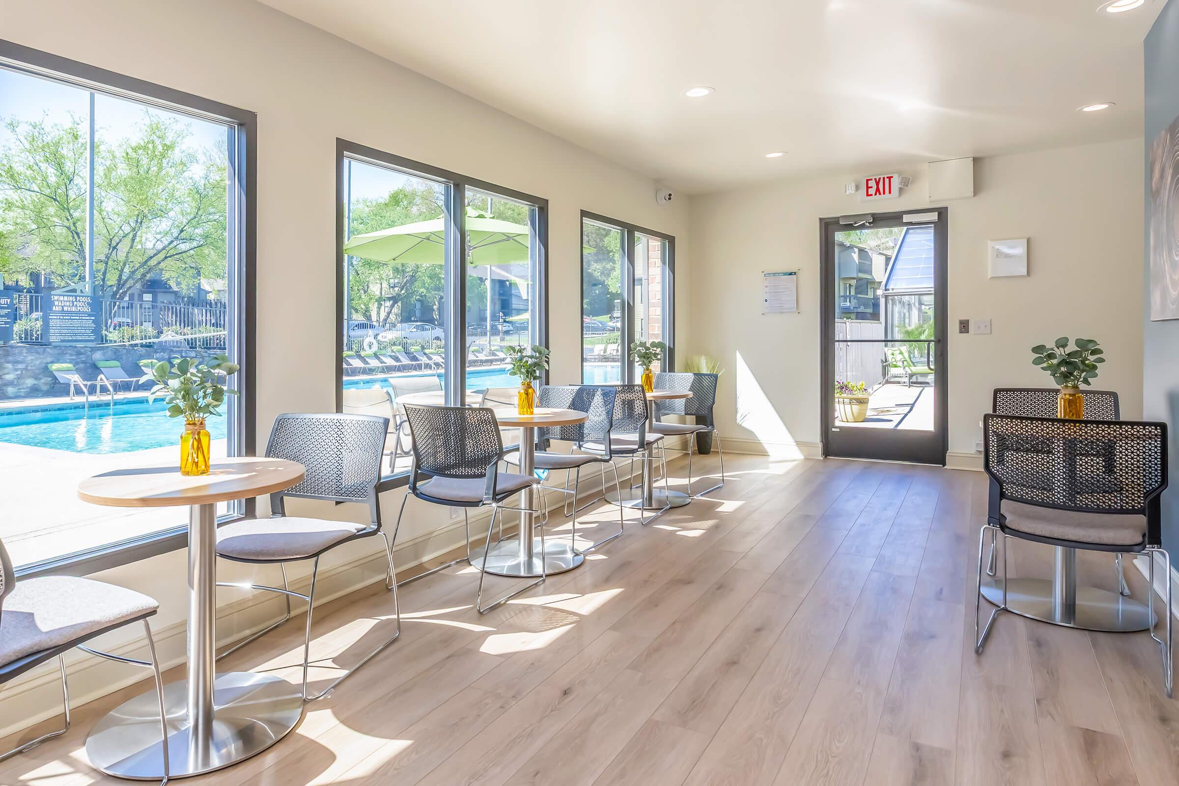 Brightly lit indoor lounge area with several small round tables and chairs. Large windows offer a view of an outdoor pool area surrounded by greenery. A few vases with plants are placed on the tables. A door leads outside, and the space has a modern, inviting feel.