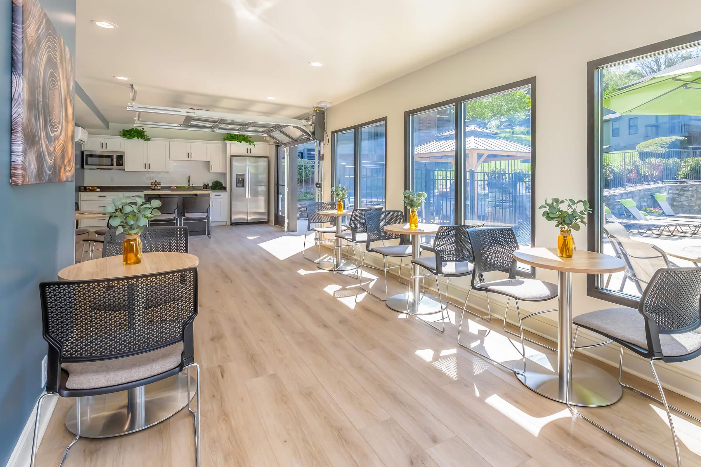 Bright, modern communal space with large windows offering natural light. Features several small tables with chairs, indoor plants in vases, and a kitchenette in the background. A garage-style door opens to an outdoor area with poolside amenities visible through the windows. Stylish and inviting atmosphere.