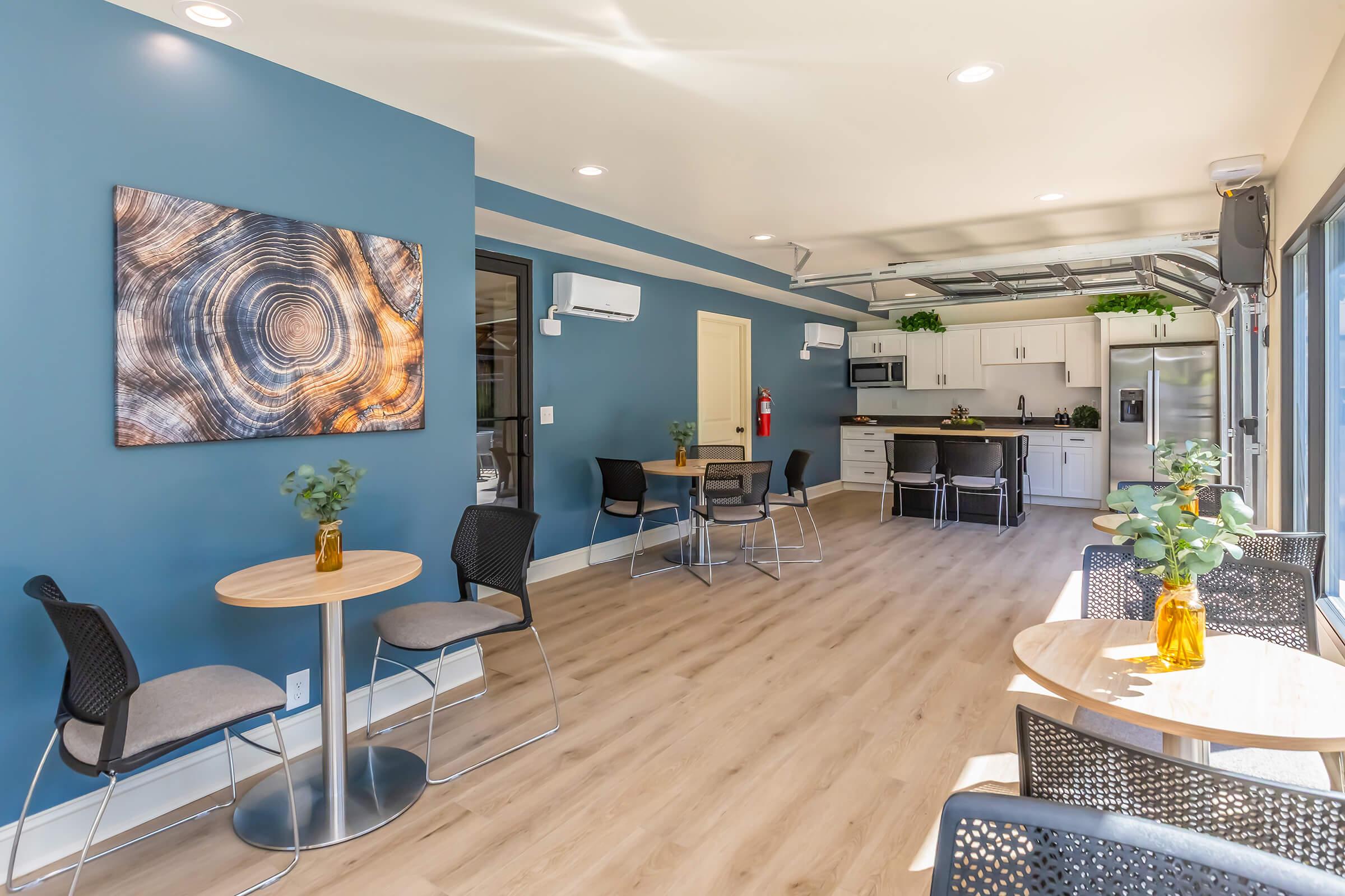 Interior view of a modern communal space featuring light wood flooring, blue walls, and a large circular wooden artwork. There are several small round tables with chairs, potted plants on the tables, and a kitchenette area in the background. The space is well-lit with natural and overhead lighting.