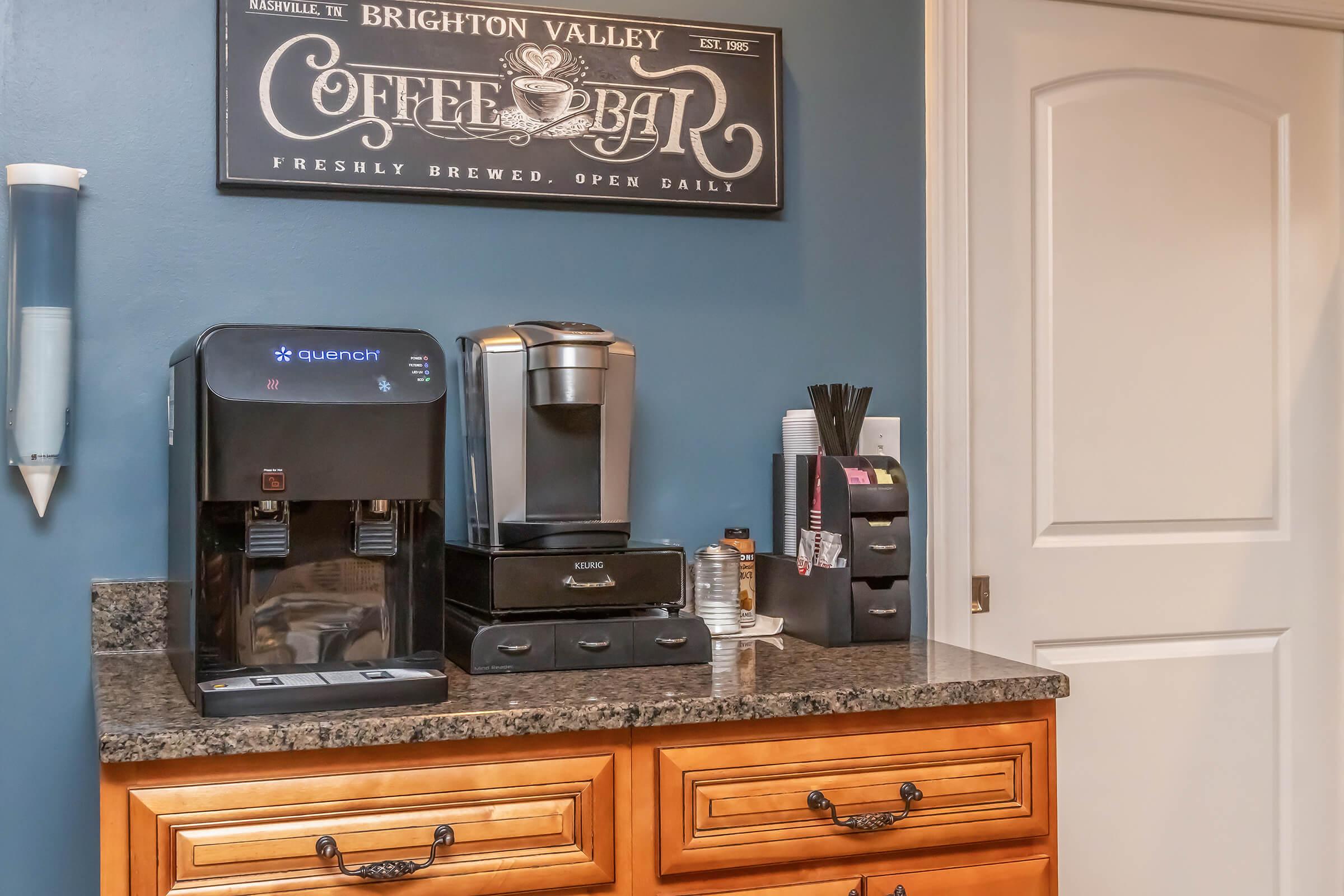 A kitchen coffee bar featuring a black coffee maker, a water dispenser labeled "Quench," and a storage unit with coffee accessories. The wall above displays a decorative sign that reads "Coffee Bar" with the establishment date 1985. The cabinetry is wooden with a smooth countertop.