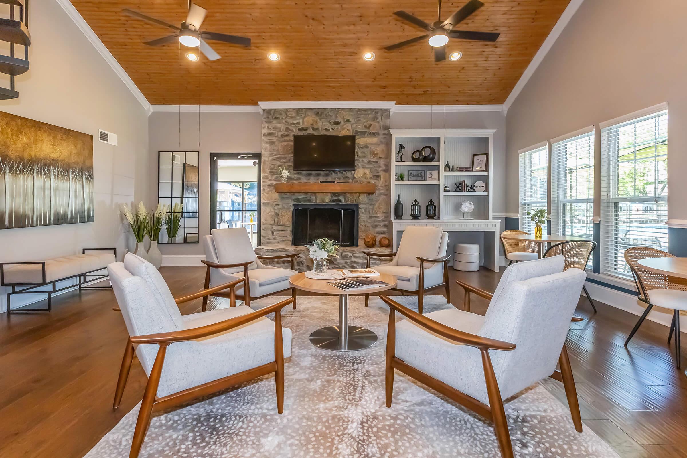 A modern living room featuring a cozy stone fireplace, wooden ceiling, and stylish furniture. It includes four armchairs around a circular table, a decorative rug, and large windows with natural light. A decorative mirror and shelves with decor items enhance the inviting atmosphere.