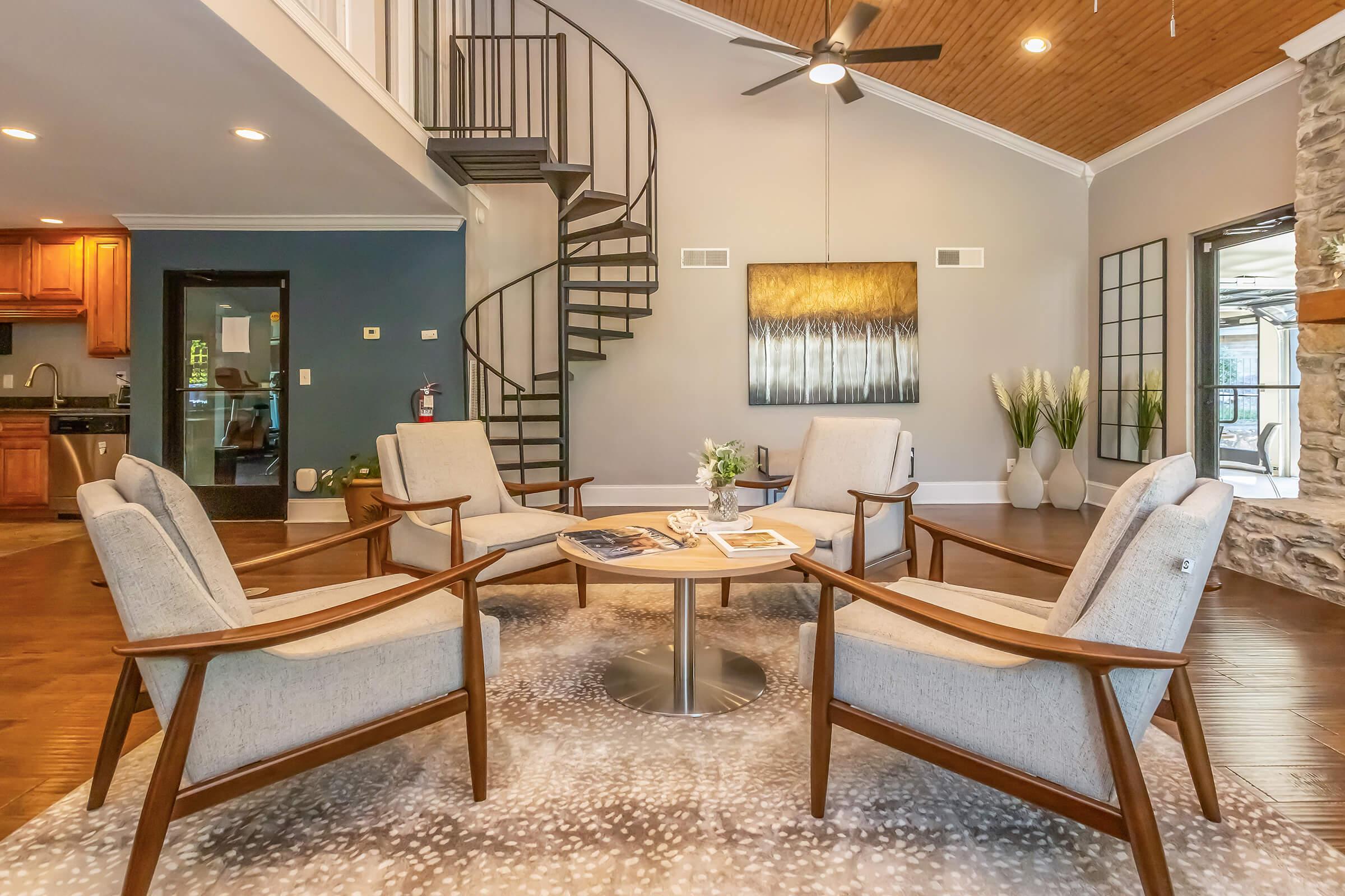 A modern living space featuring a circular seating arrangement with four armchairs around a small table, a spiral staircase in the background, and a cozy atmosphere enhanced by natural light and stylish decor, including a decorative painting and plants.