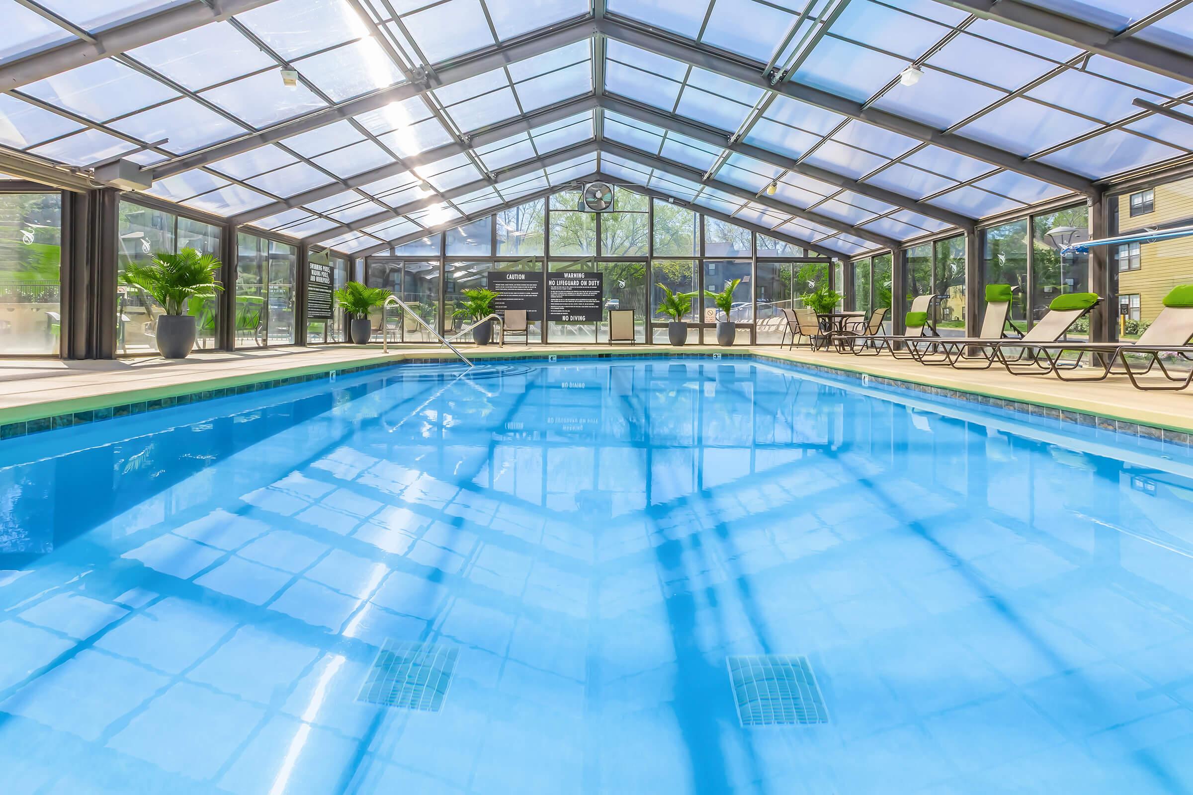 Indoor swimming pool with a clear blue water under a glass roof. Surrounding the pool are lounge chairs and potted plants. Bright natural light filters in, reflecting off the water's surface. The atmosphere is serene and inviting, perfect for relaxation and leisure activities.