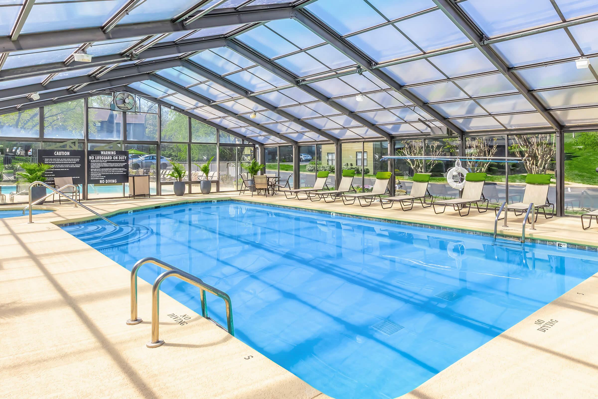 Indoor swimming pool with a transparent roof, surrounded by lounge chairs. The clean blue water is inviting, and large windows allow natural light to fill the space. The atmosphere is bright and airy, perfect for relaxation and leisure.