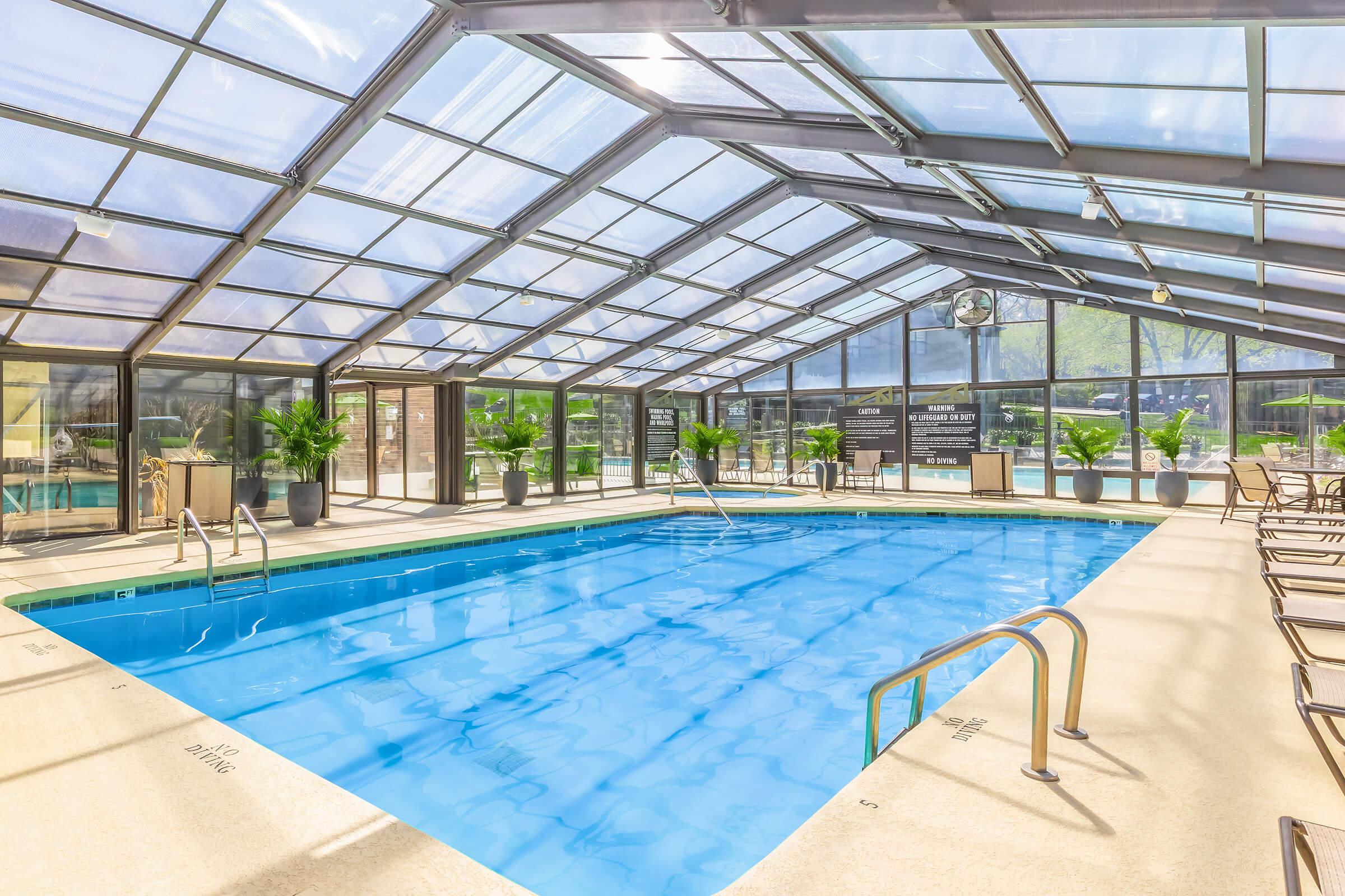 Indoor swimming pool area with a clear, glass ceiling allowing natural light. The pool features ladders at both ends and is surrounded by potted plants and lounge chairs along the perimeter. The space is bright and inviting, designed for relaxation and leisure.