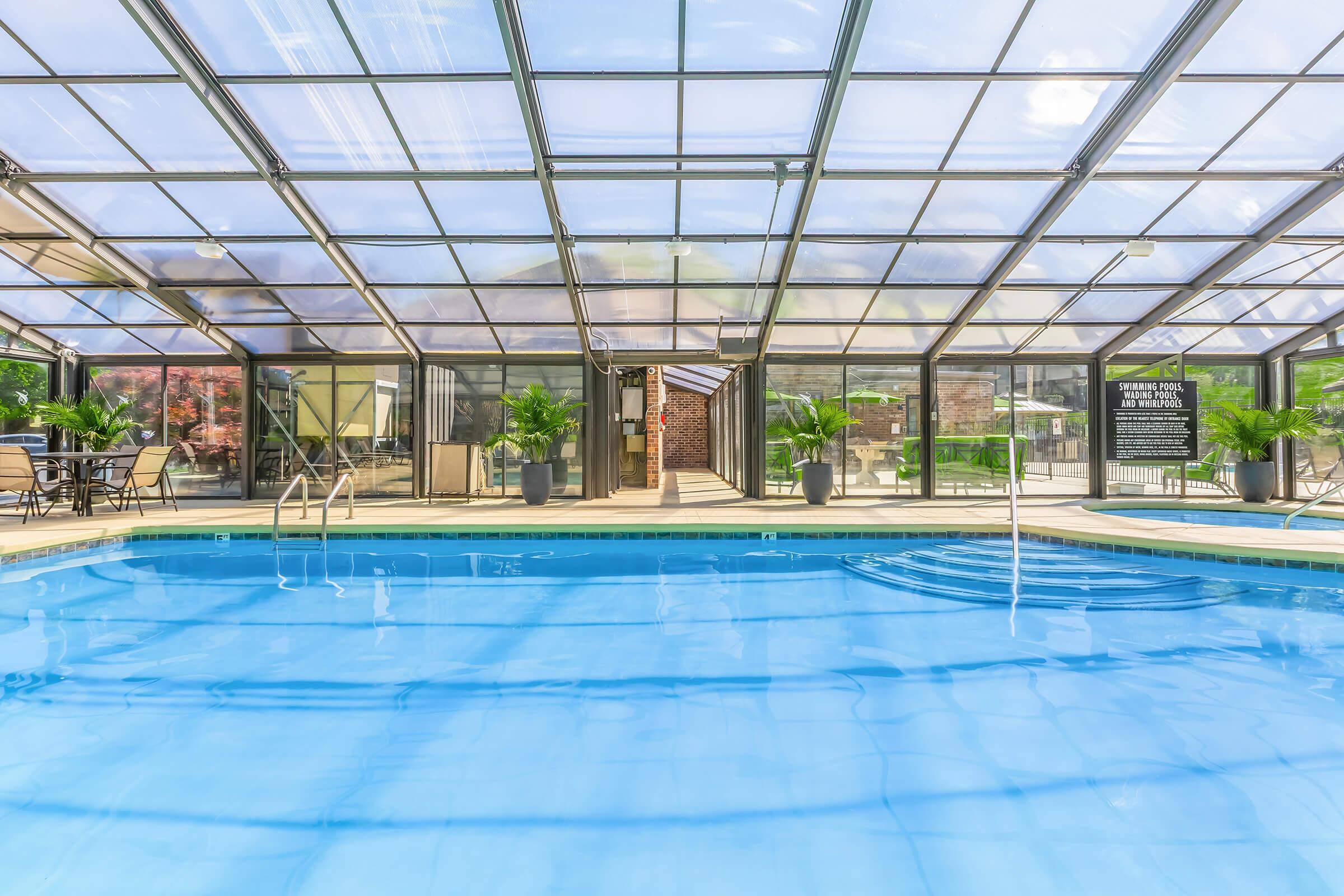 Indoor swimming pool with clear, vaulted glass ceiling. The pool water is a bright blue, and there are potted plants around the area. Lounge chairs are set up nearby, and there is a sign on the wall in the background, indicating rules or information about the facility. The atmosphere is bright and inviting.
