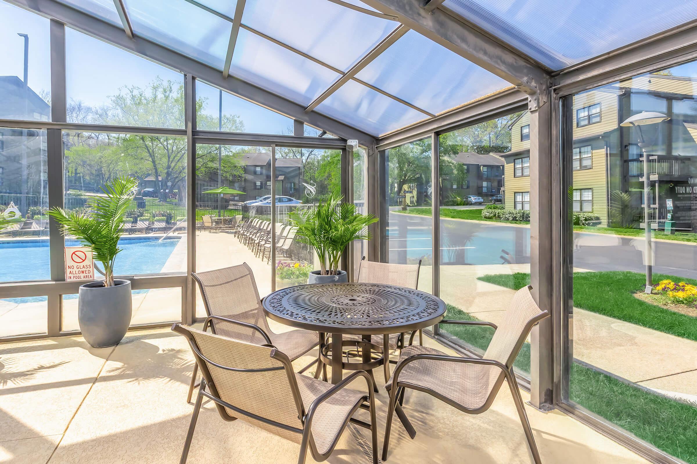 A sunlit indoor seating area featuring a round metal table surrounded by four chairs, with potted plants and a view of a pool area outside. Large glass windows provide a bright atmosphere, while a sign indicates no diving in the pool. The setting is inviting and relaxing, ideal for social gatherings.