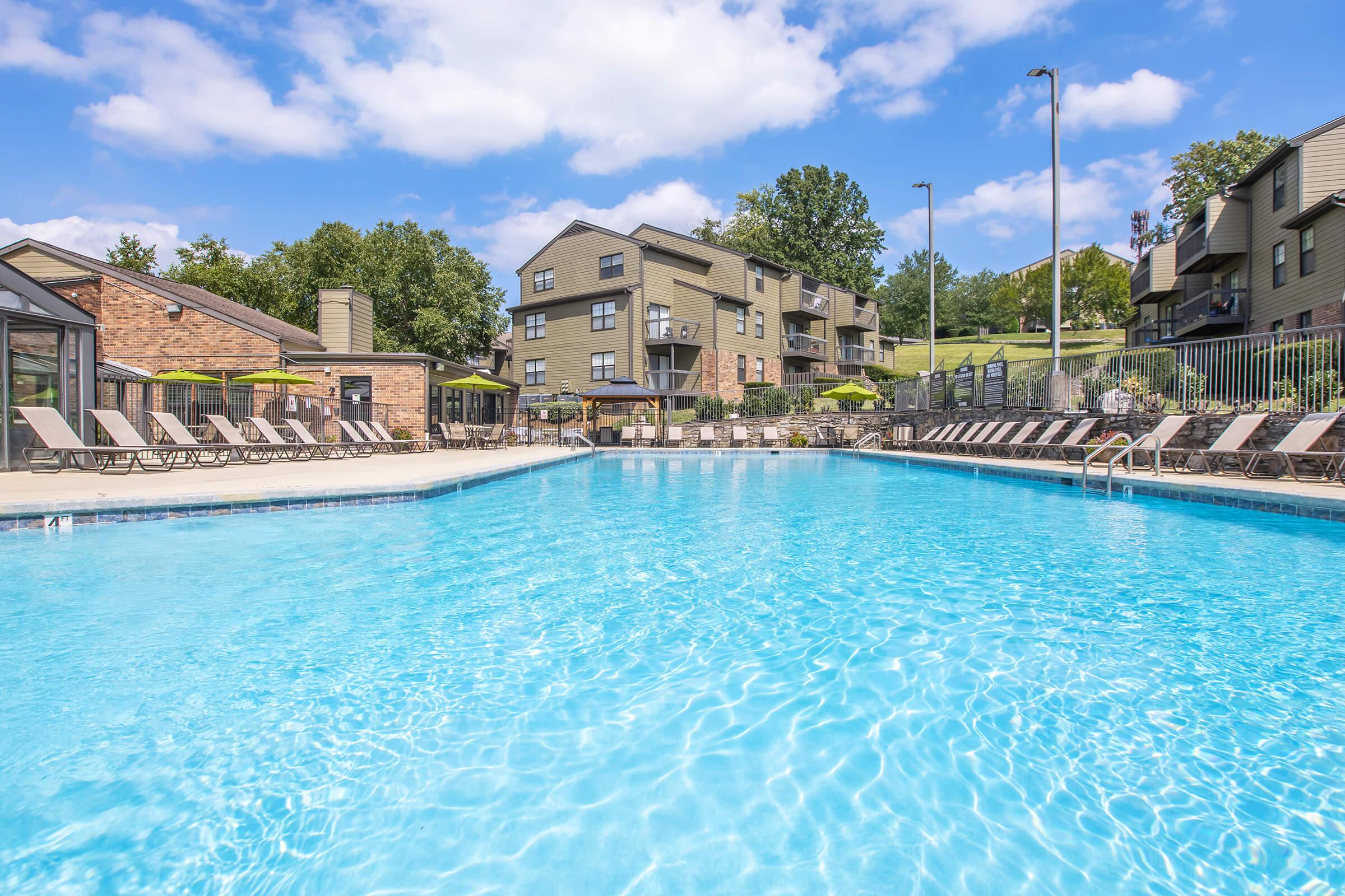 A clear, inviting swimming pool with blue water reflecting the sky, surrounded by lounge chairs. In the background, there are several modern apartment buildings and lush greenery. The scene is bright and sunny, suggesting a relaxing outdoor space.