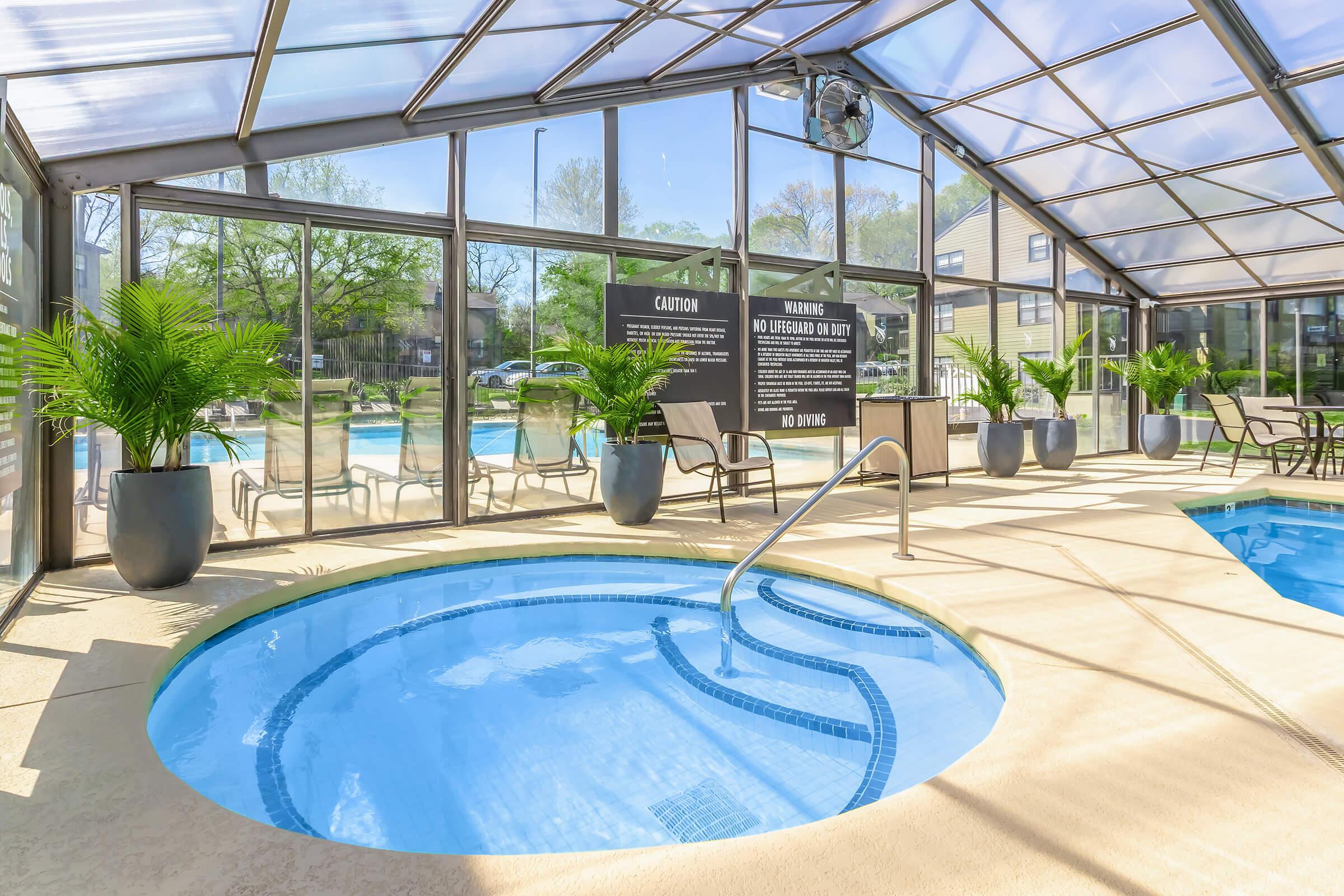 Indoor pool area with a hot tub, surrounded by large glass panels that create a bright atmosphere. Potted plants are placed around the space, and there are chairs for seating. A caution sign is visible, along with a "No Diving" warning. The background shows a larger swimming pool outside.