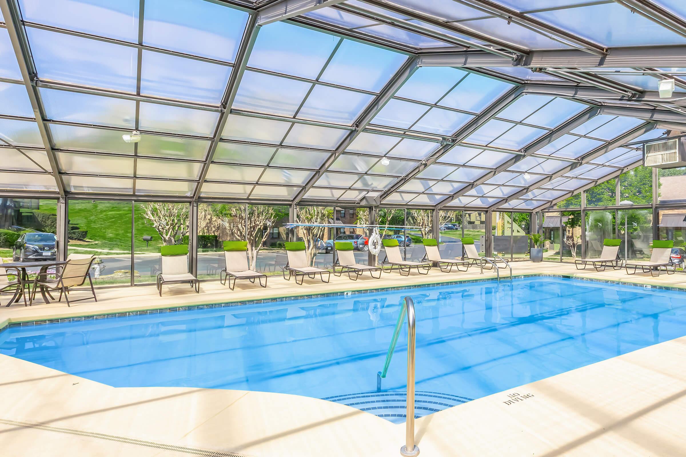 Indoor swimming pool with a glass ceiling, surrounded by lounge chairs. The area is well-lit, featuring greenery outside the large windows. Steps lead into the pool, which has clear blue water, and the space appears clean and inviting for relaxation or exercise.