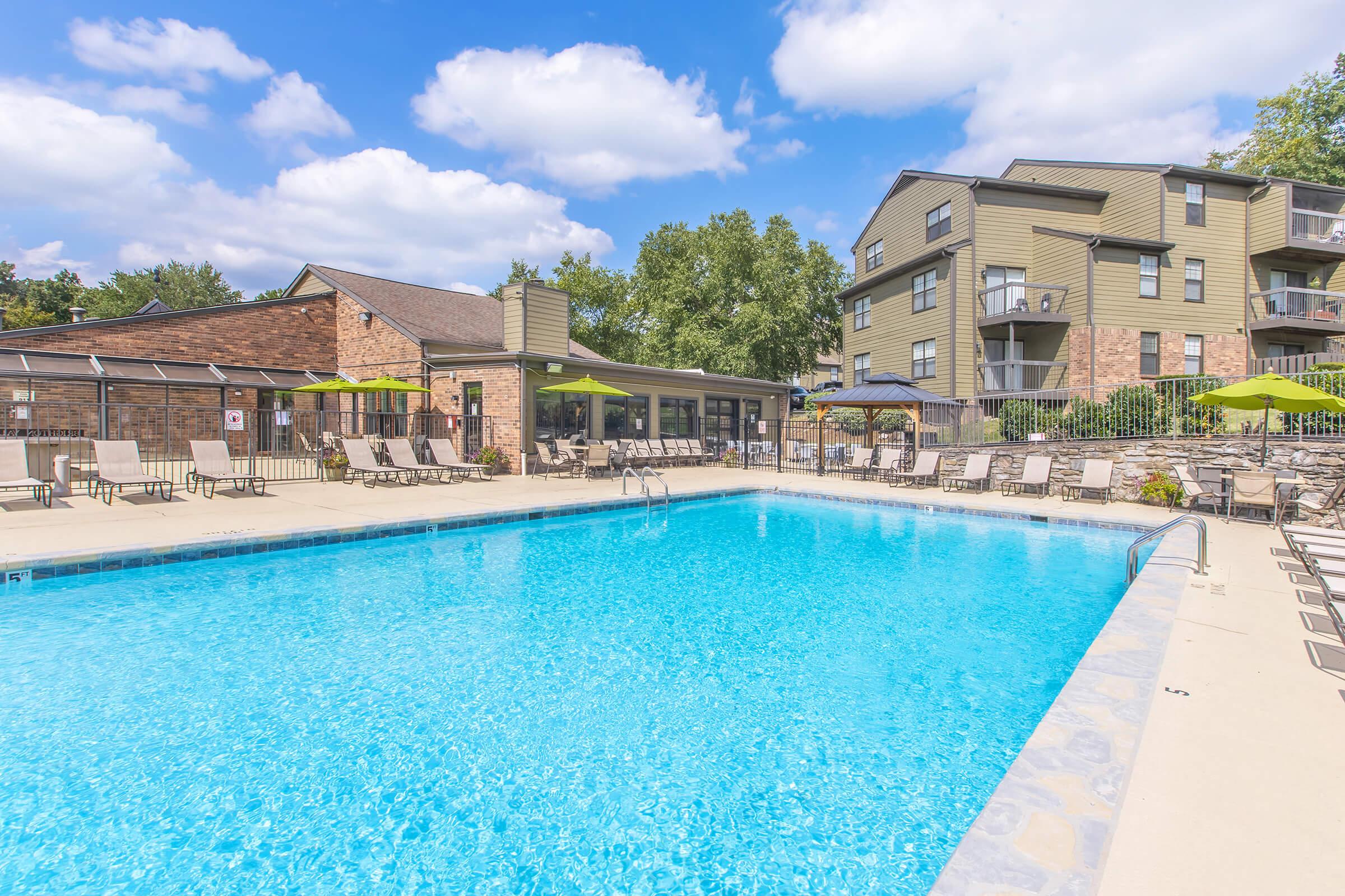 A clear blue swimming pool surrounded by lounge chairs and tables, with green umbrellas providing shade. In the background, there are modern apartment buildings and a shaded patio area. The sky is partly cloudy, creating a bright and inviting atmosphere.