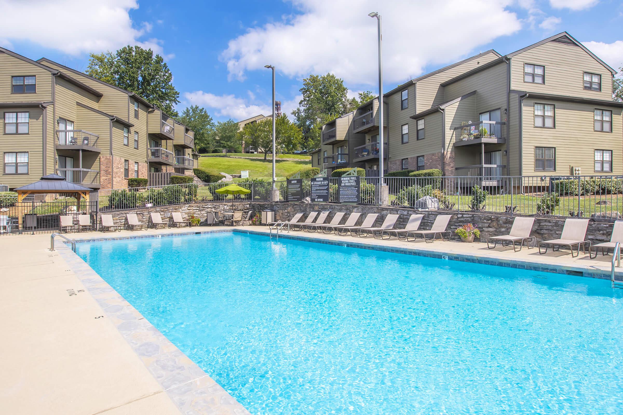 A large outdoor swimming pool surrounded by lounge chairs, with several apartment buildings visible in the background. The scene is set on a sunny day with blue skies and green landscaping, creating a relaxed and inviting atmosphere for residents and guests.
