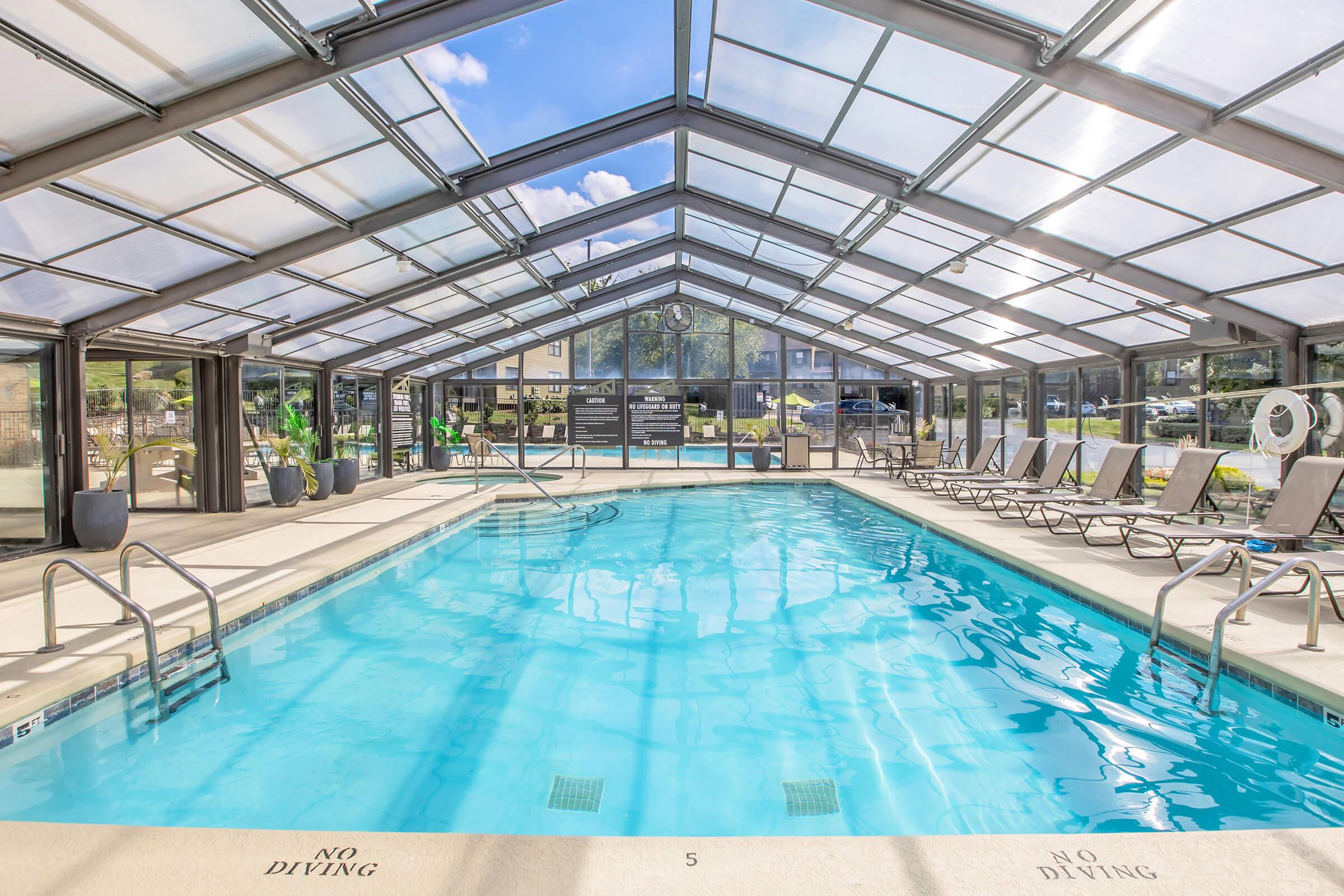 Indoor swimming pool with a transparent roof, surrounded by lounge chairs. Natural light fills the space, highlighting clear blue water. Signs indicate pool rules, while large windows provide views of the outdoors. The setting is clean and modern, ideal for relaxation and recreation.
