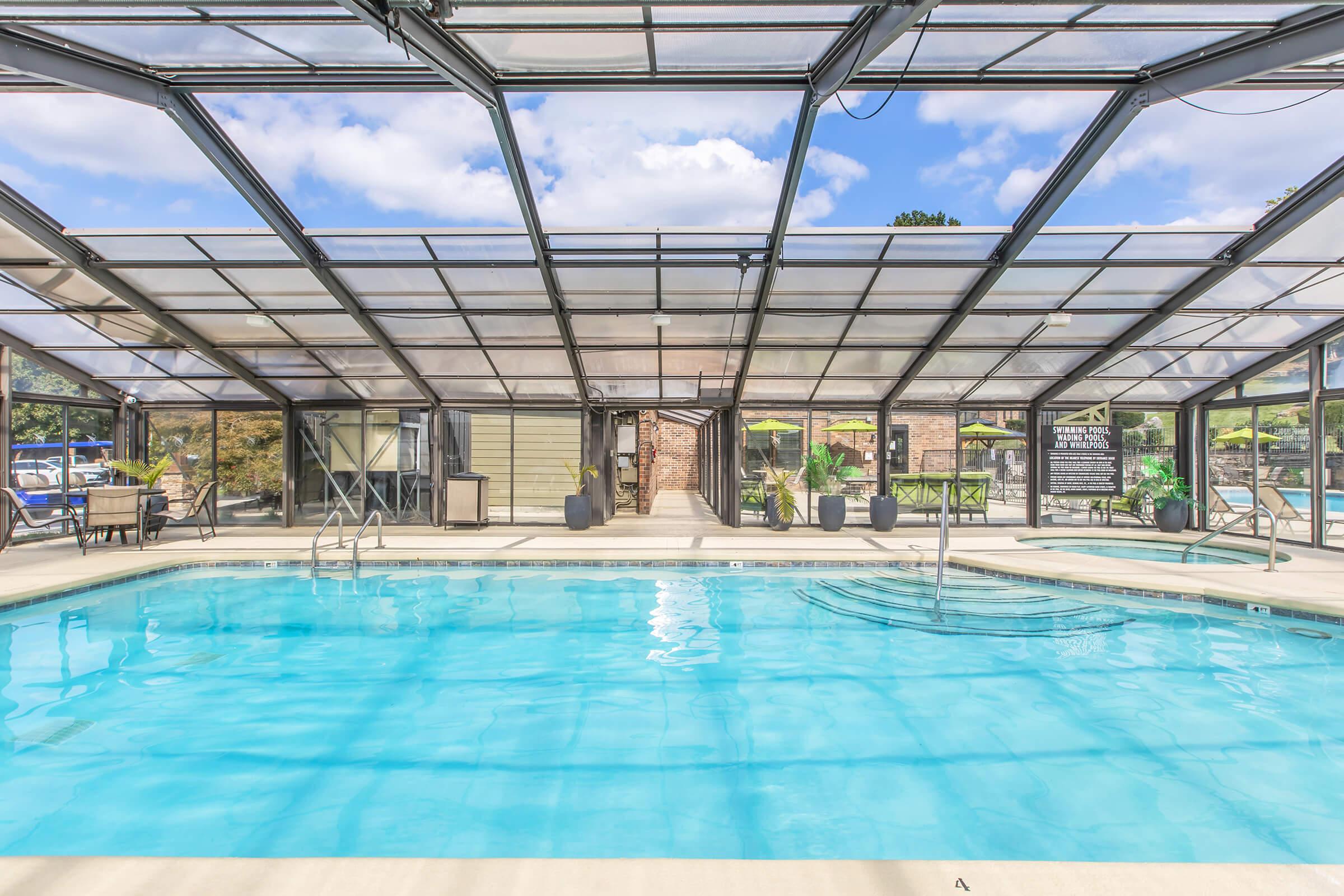Indoor swimming pool surrounded by large glass panels, allowing natural light to fill the space. The pool is clear and inviting, with steps leading in. Potted plants and lounge chairs are positioned around the pool area, creating a relaxing atmosphere. The background features a wooden deck and additional outdoor seating.