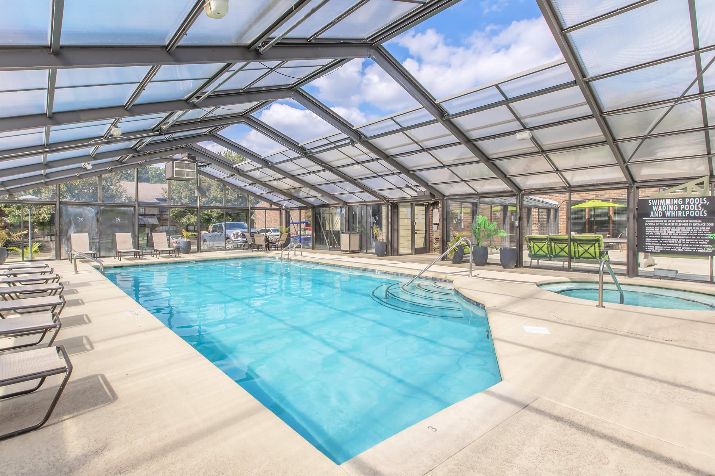 Indoor swimming pool area with a clear glass roof, featuring a spacious pool, whirlpool, and lounge chairs. Sunlight streams in, creating a bright atmosphere. In the background, there are green plants and signage detailing facilities. Ideal for relaxation and leisure.