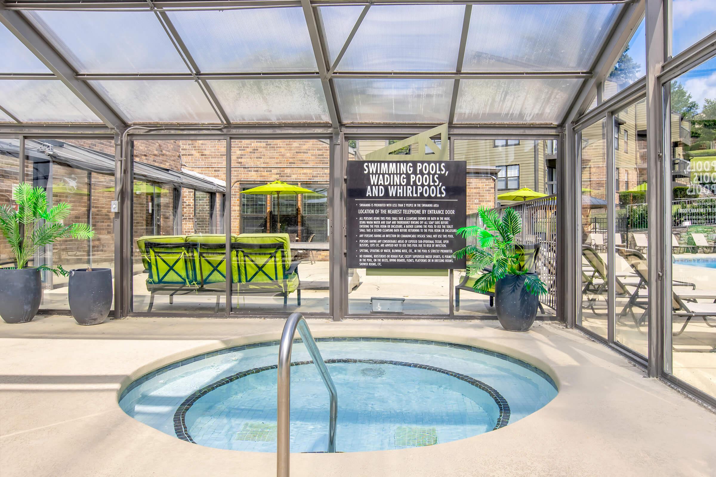 Indoor area featuring a whirlpool with steps, surrounded by a patio space. The background includes lounge chairs and tables under umbrellas. A sign on the wall mentions "Swimming Pools, Wading Pools, and Whirlpools." Green potted plants add a touch of greenery to the space.