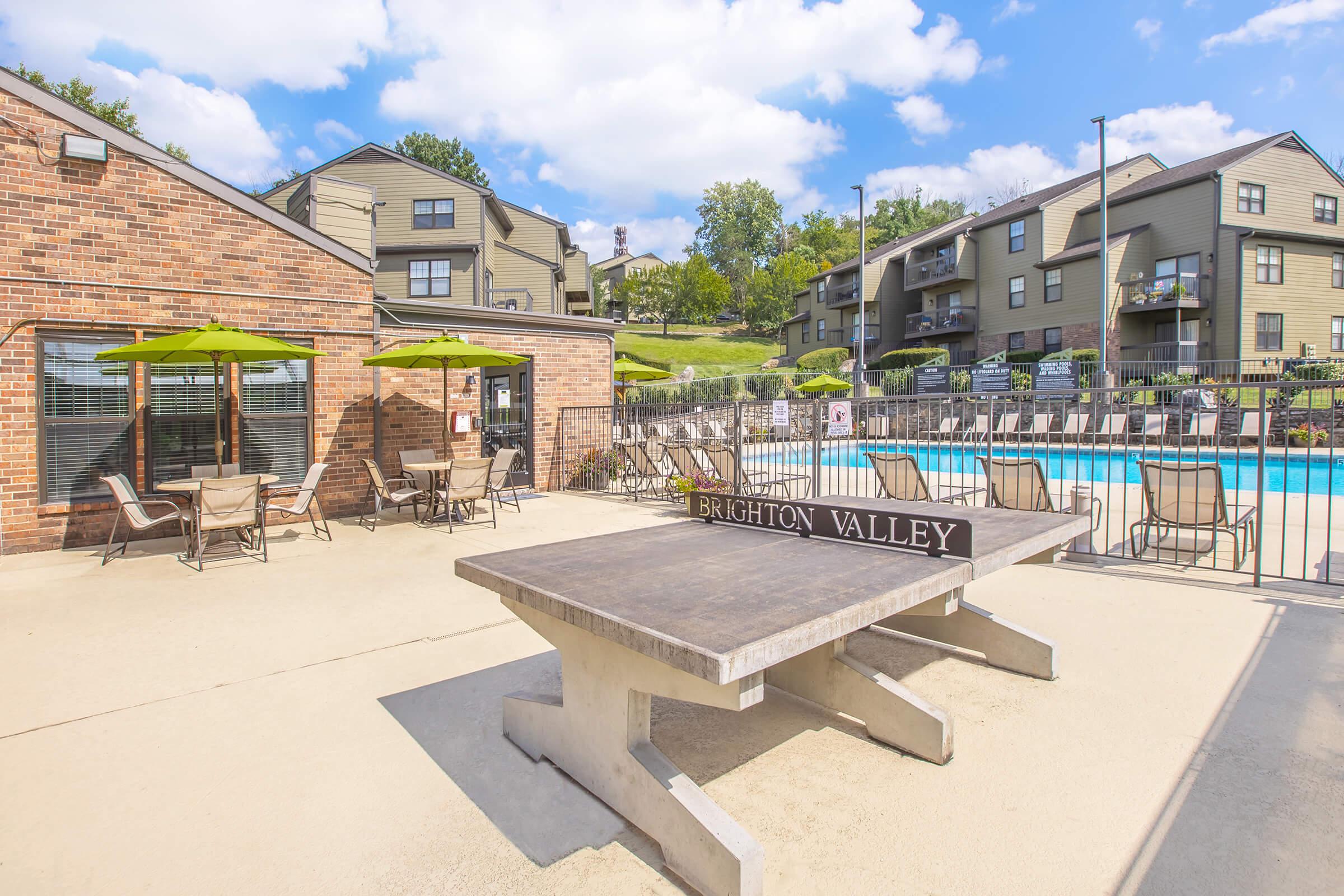 A view of an outdoor pool area at Brighton Valley, featuring a table and chairs under green umbrellas, with an apartment building in the background. The pool is adorned with lounge chairs, and the scene is set against a clear blue sky and green landscaping.